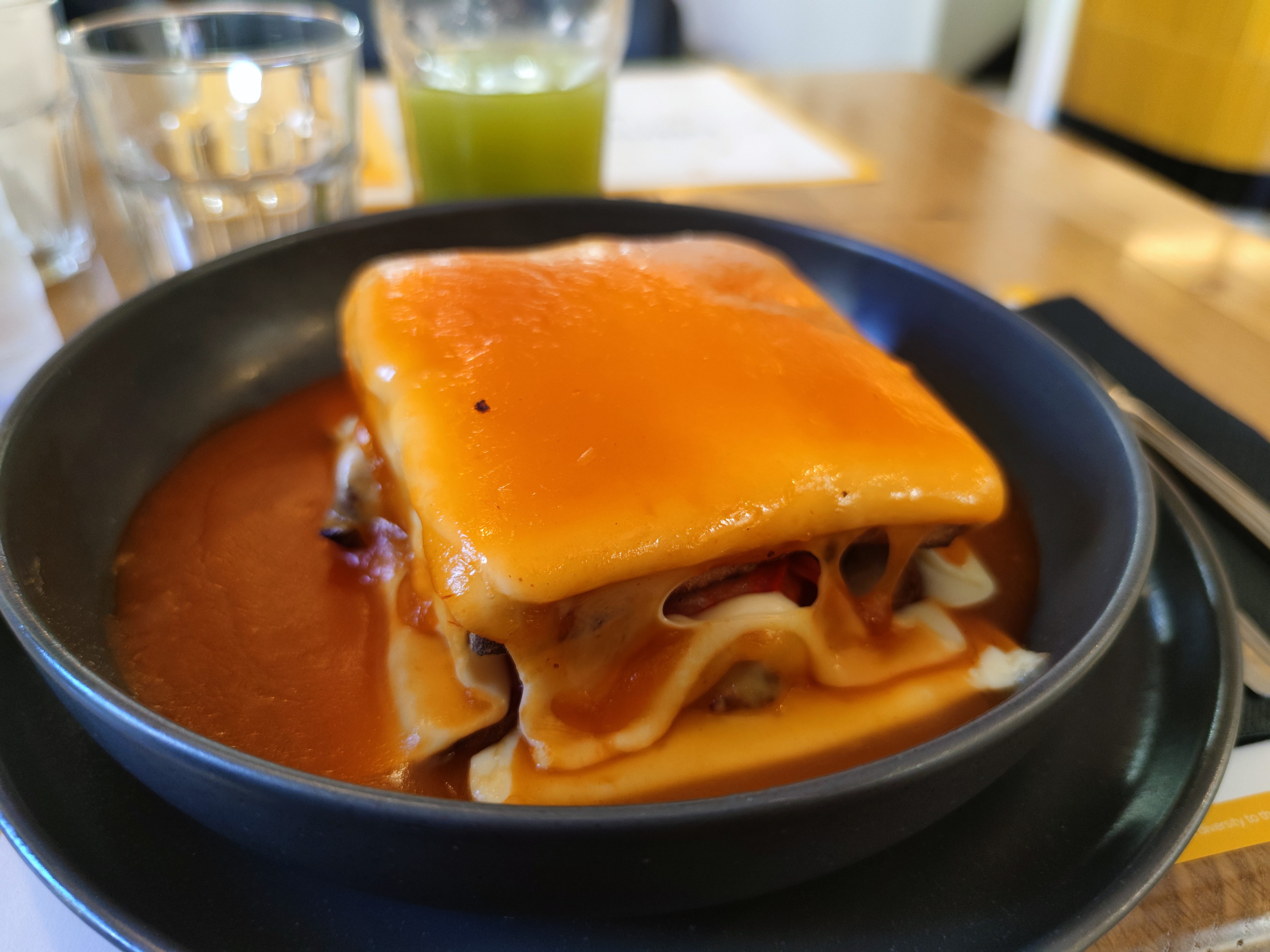A close-up shot of a Portuguese Francesinha sandwich served in a dark, round bowl. The square sandwich is completely covered in a thick layer of smooth, melted yellow cheese and sits in a pool of rich, orange-red tomato and beer sauce.