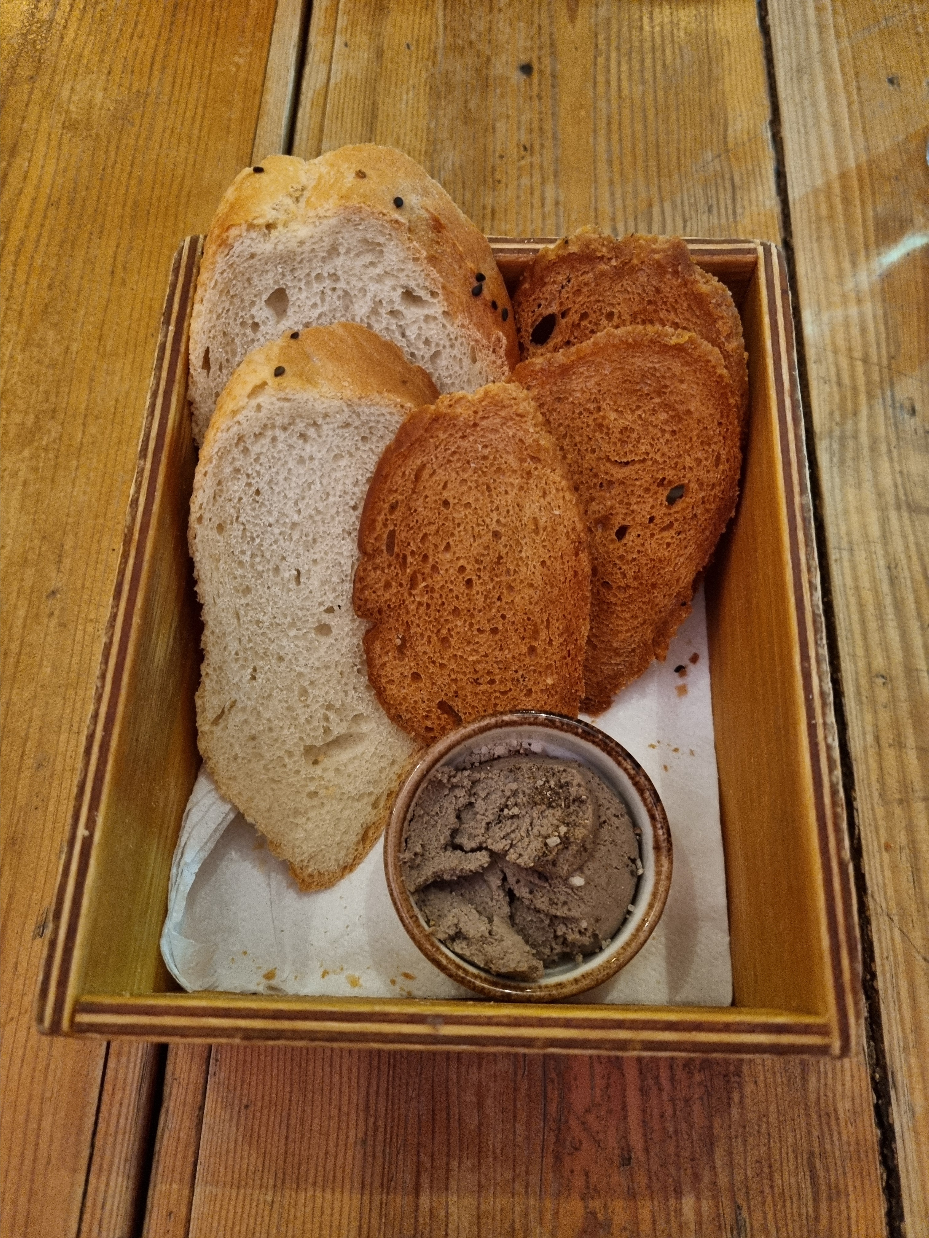A rectangular wooden tray holds an appetizer featuring three thick slices of soft, light-colored bread with a crust, three pieces of crispy, dark-fried bread, and a small ceramic bowl of duck offal and port wine pate.