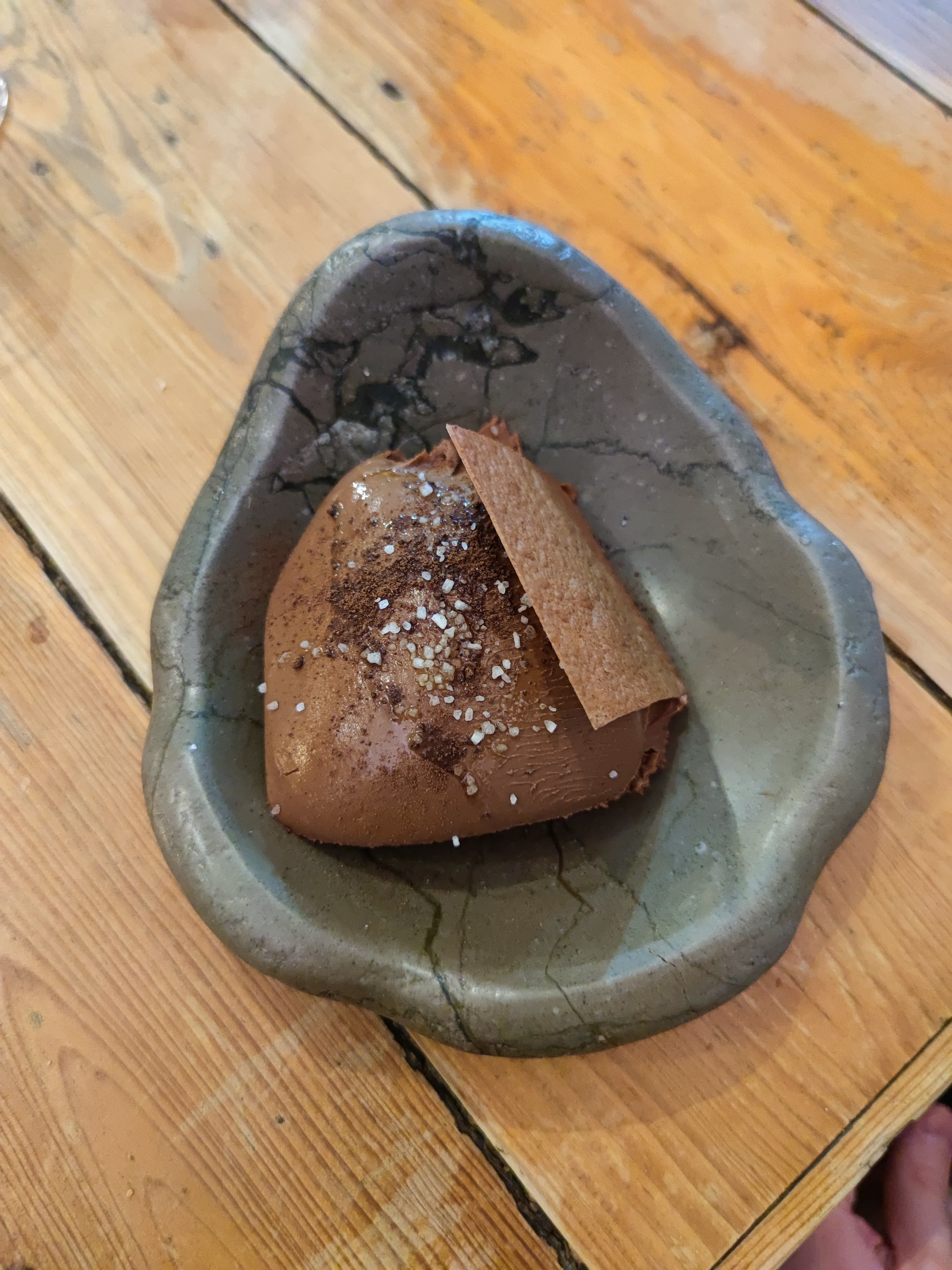 A close-up of the 'Chocolate, Olive Oil and Salt' dessert from Oficina do Duque, presented in a distinctive, irregular-shaped dark stone-like bowl. A large, smooth mound of rich brown chocolate mousse is topped with visible salt crystals and chocolate shavings, accompanied by a thin, rectangular, reddish-brown crisp.