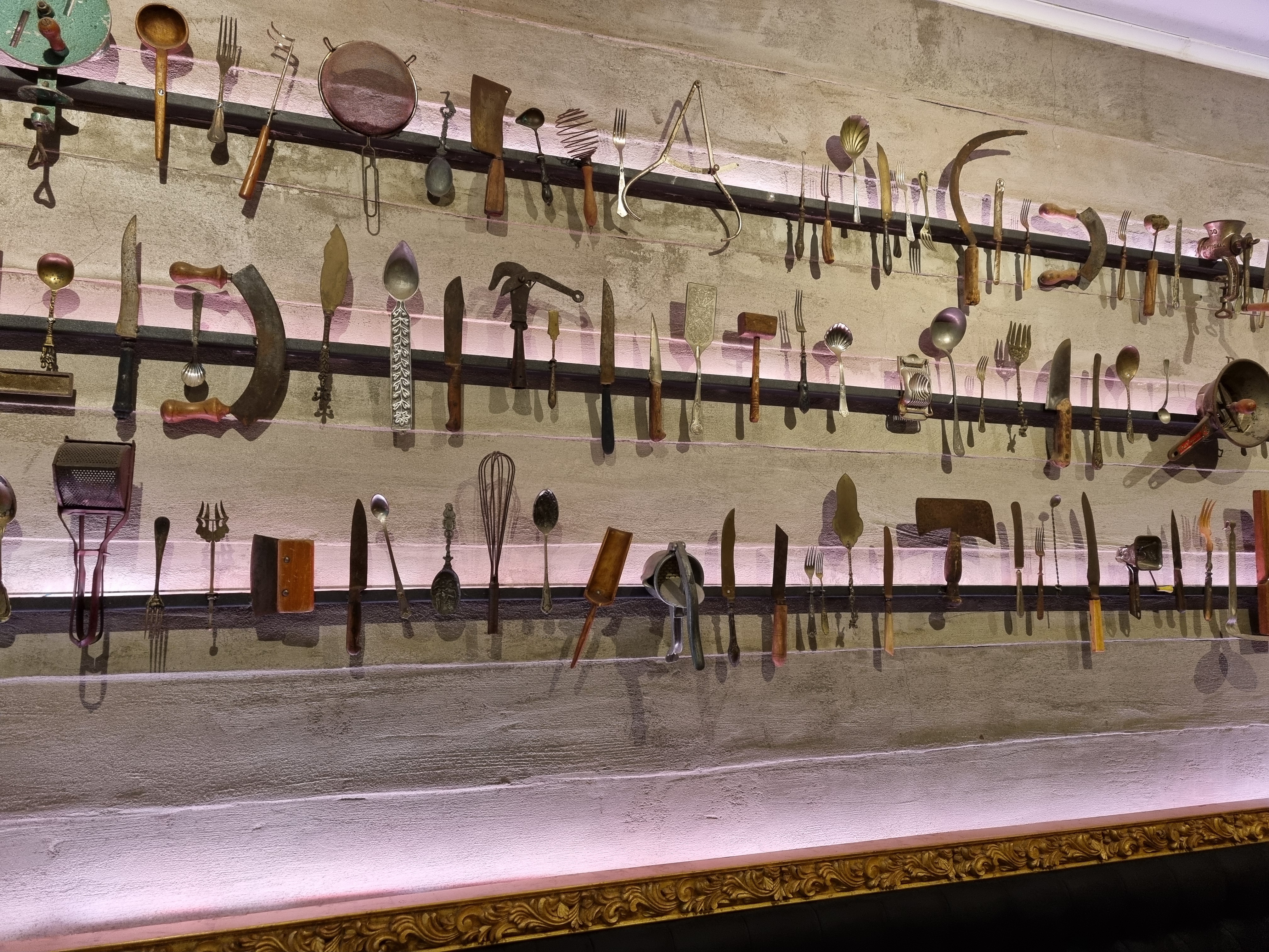 A close-up view of a restaurant's wall, featuring a unique decorative display of numerous vintage kitchen implements like knives, forks, whisks, and other cooking tools arranged neatly in horizontal rows against a light-colored background.