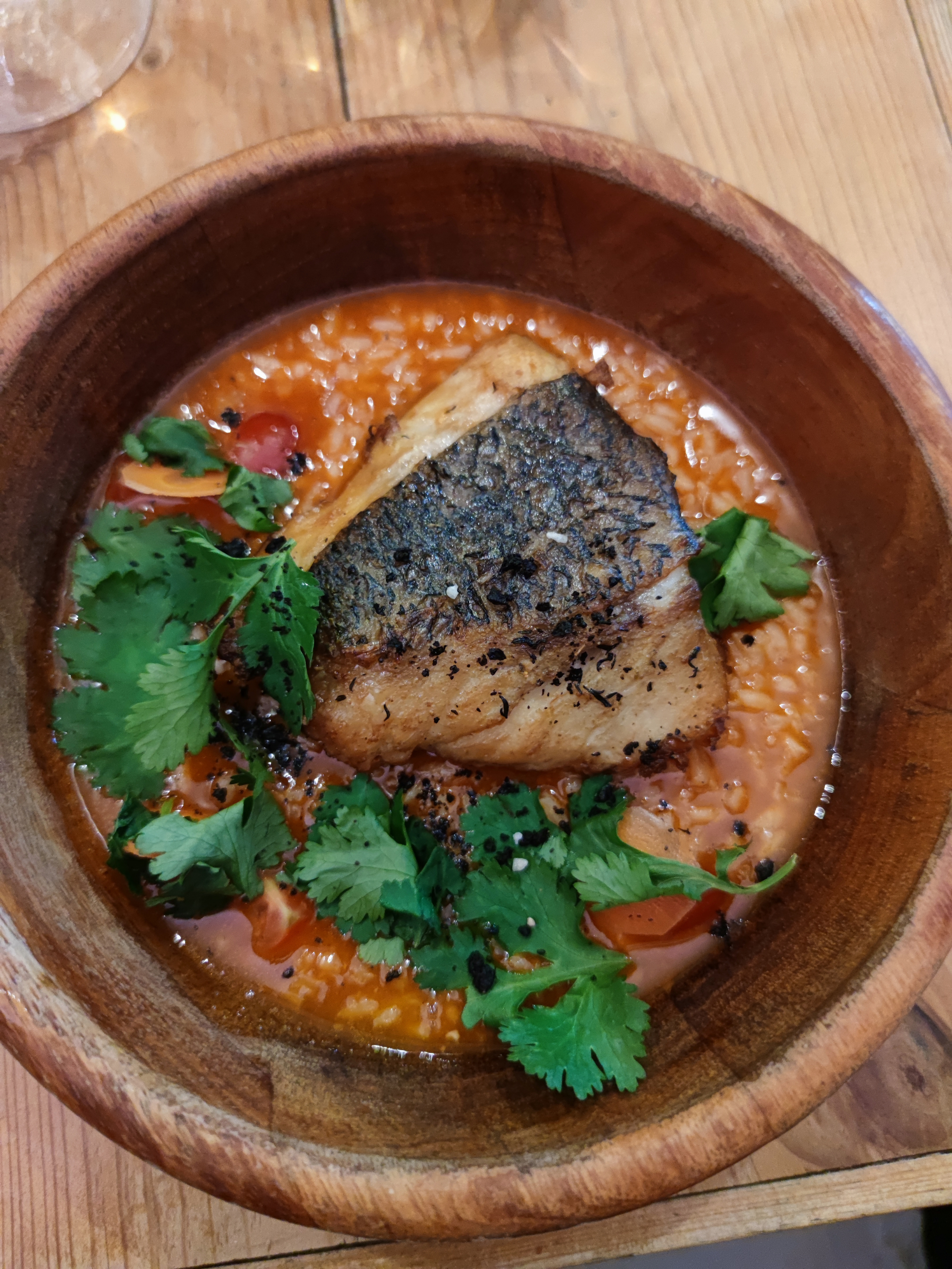 A close-up of the 'Meagre, Rice and Coriander' dish from Oficina do Duque, presented in a rustic wooden bowl. A golden-brown piece of meagre fish with speckled, crispy skin sits atop a rich, soupy tomato rice, garnished with vibrant green cilantro leaves and small black and red spices.