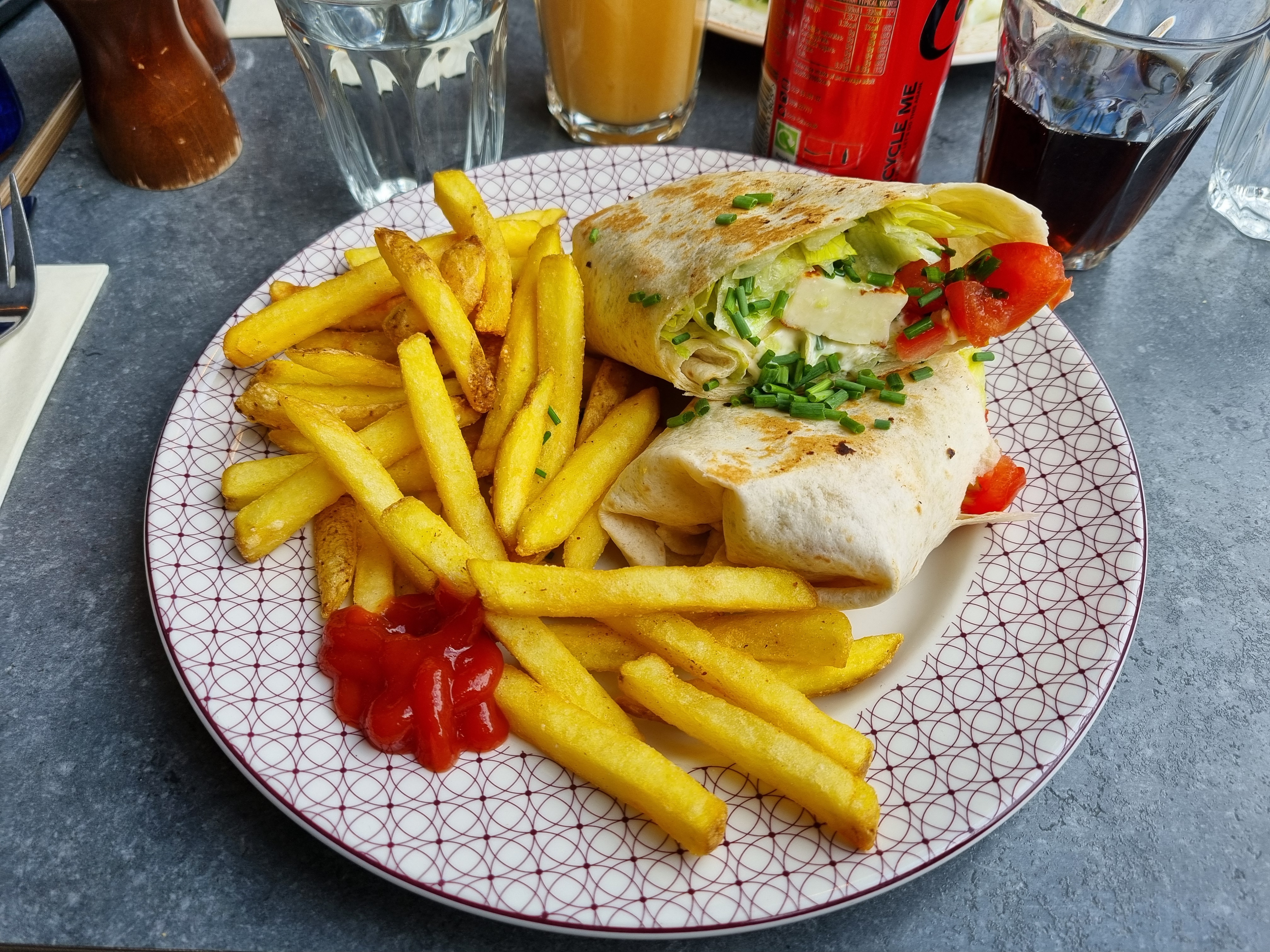 A Halloumi Wrap cut in half, revealing its fresh ingredients, served with a generous portion of golden french fries and a dollop of ketchup on a patterned white plate.