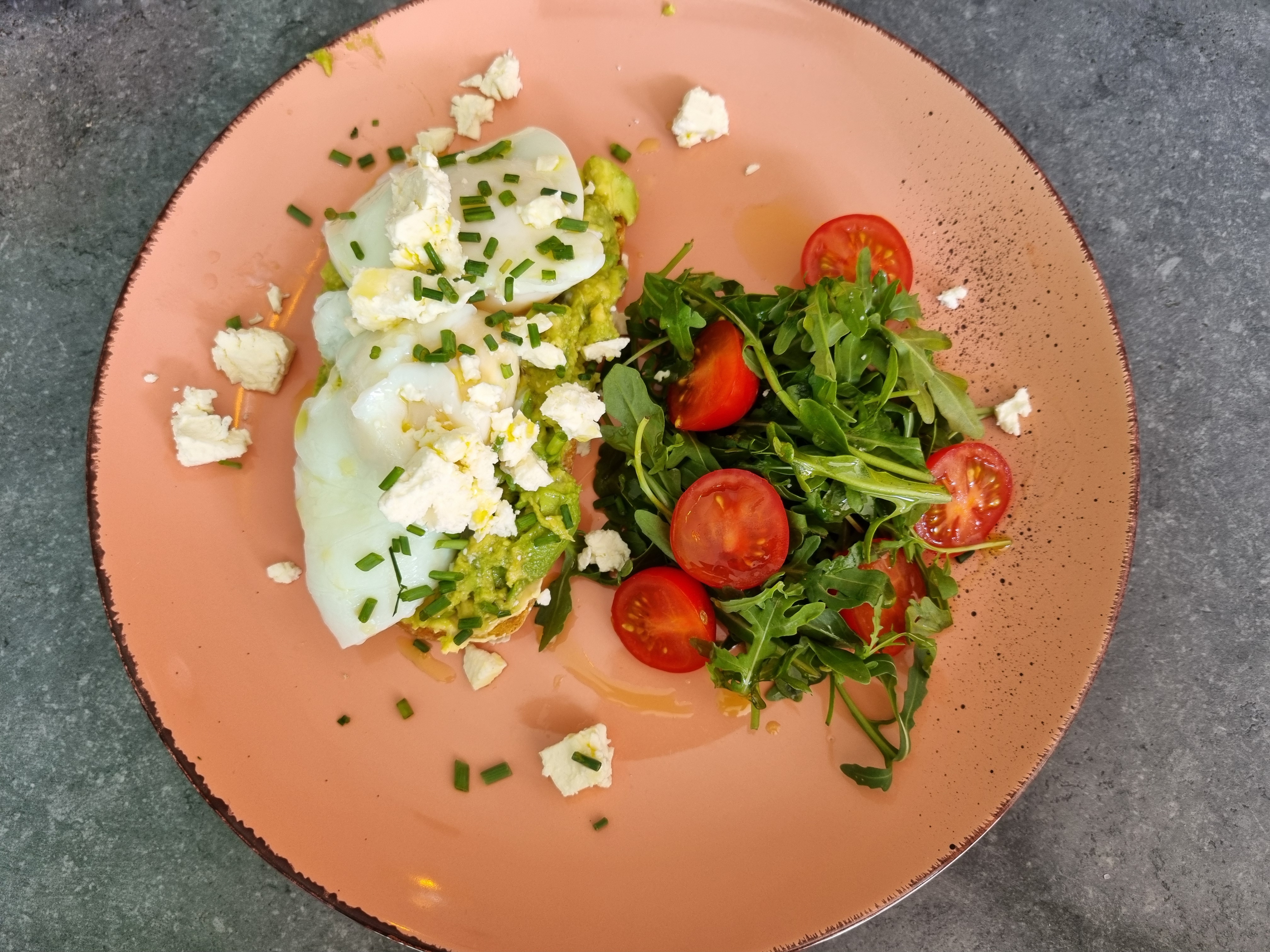 A close-up overhead shot of Redmoon Toast with perfectly poached eggs, creamy avocado spread, crumbled feta, and a side of fresh arugula salad with cherry tomatoes, all on a coral-colored plate.