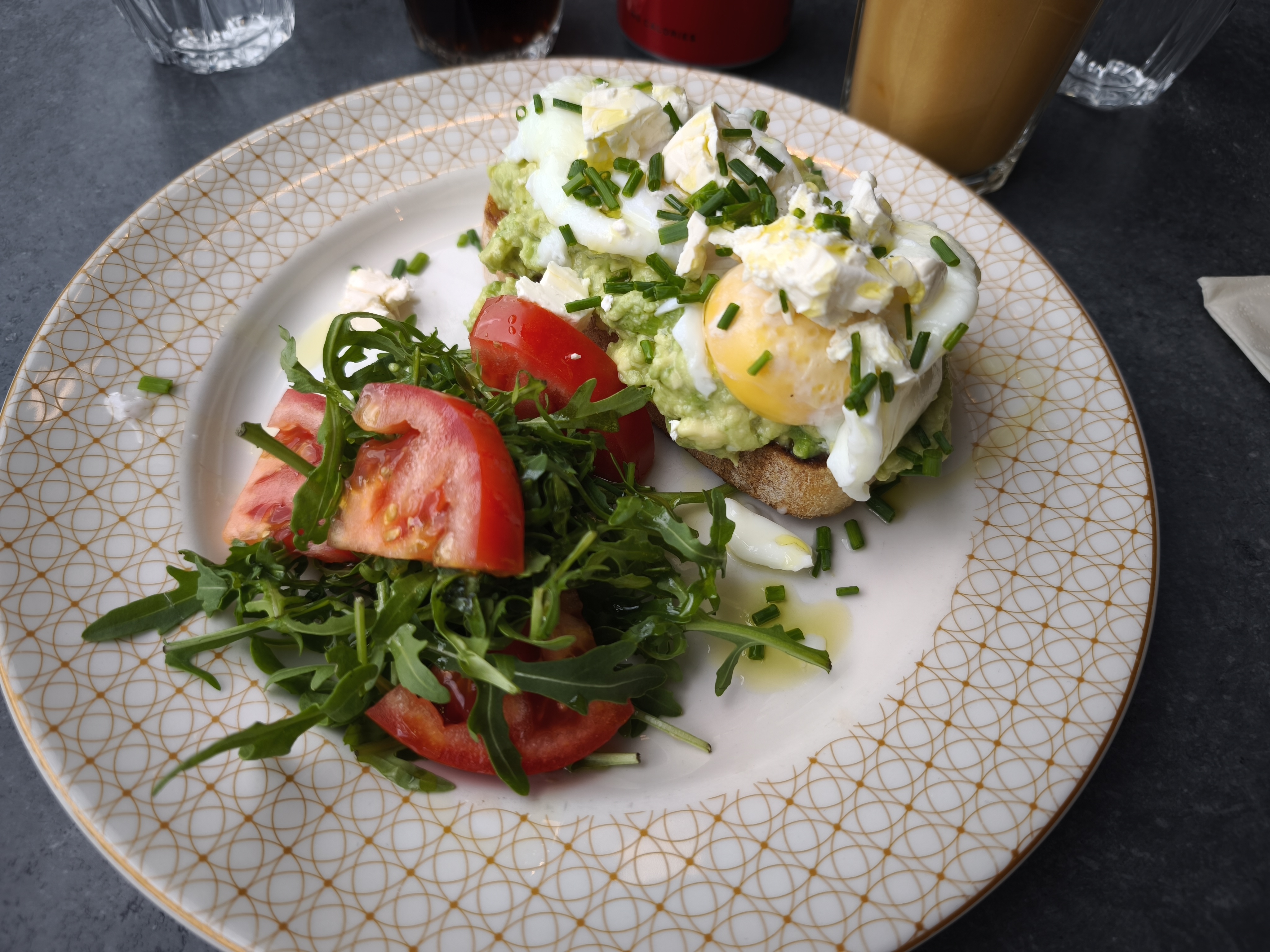 A side view of Redmoon Toast on a patterned white plate, featuring two slices of toast topped with mashed avocado, poached eggs, feta cheese, and chives, alongside a fresh green salad with sliced tomatoes.