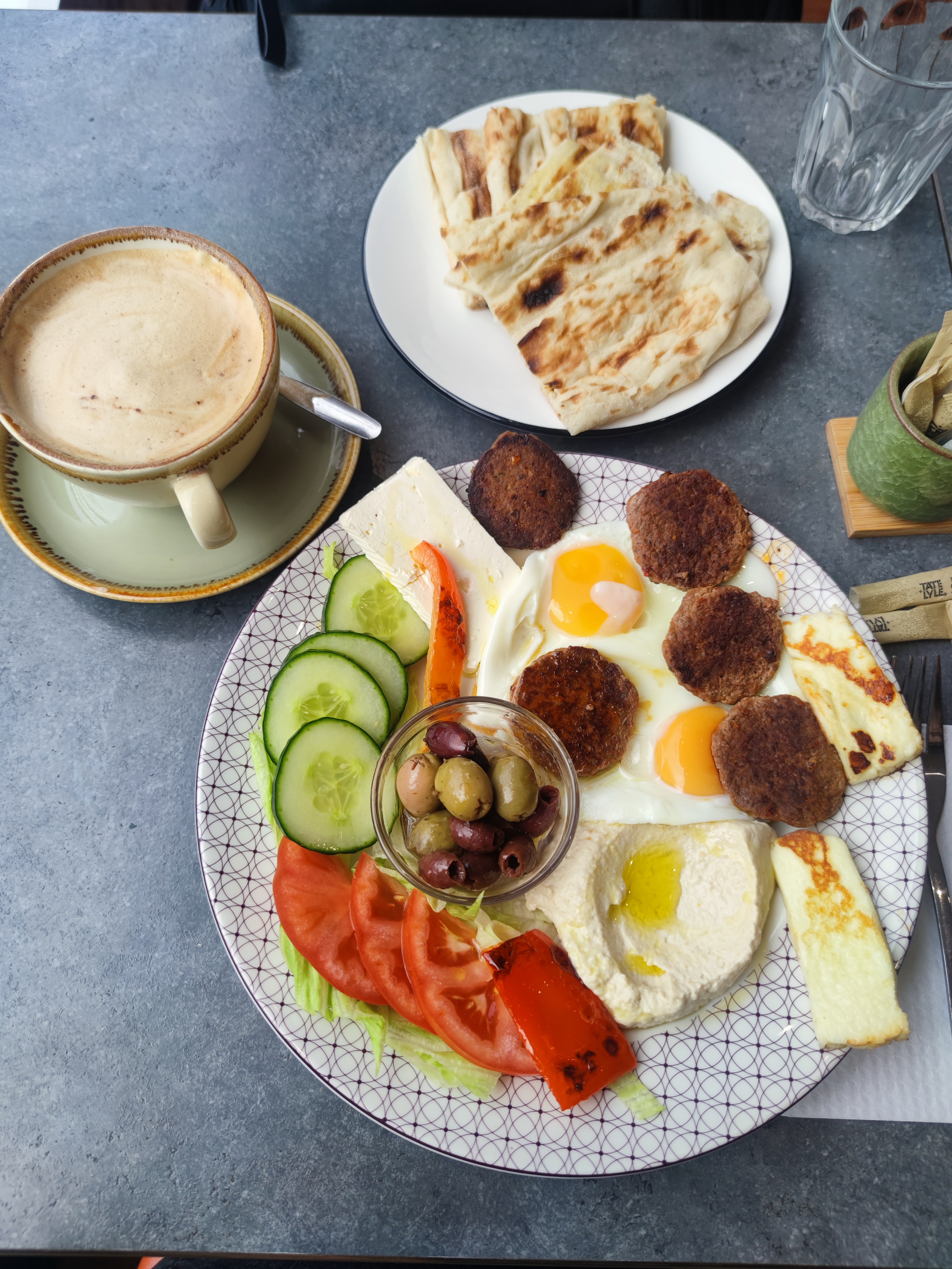 A comprehensive Turkish breakfast spread featuring fried eggs, spicy sausage patties, halloumi, hummus, fresh vegetables like cucumber and tomato, olives, and flatbread, with a coffee in the background.