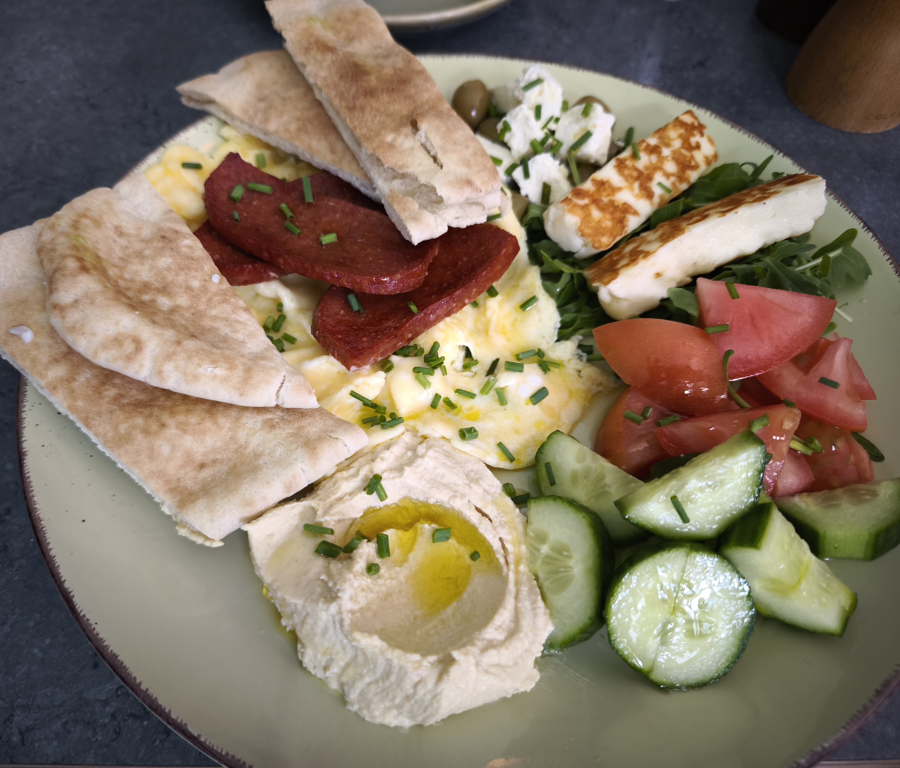 A close-up of a Turkish breakfast plate with scrambled eggs, spicy sausage, grilled halloumi, creamy hummus, fresh cucumber and tomato, feta, olives, and warm flatbread.