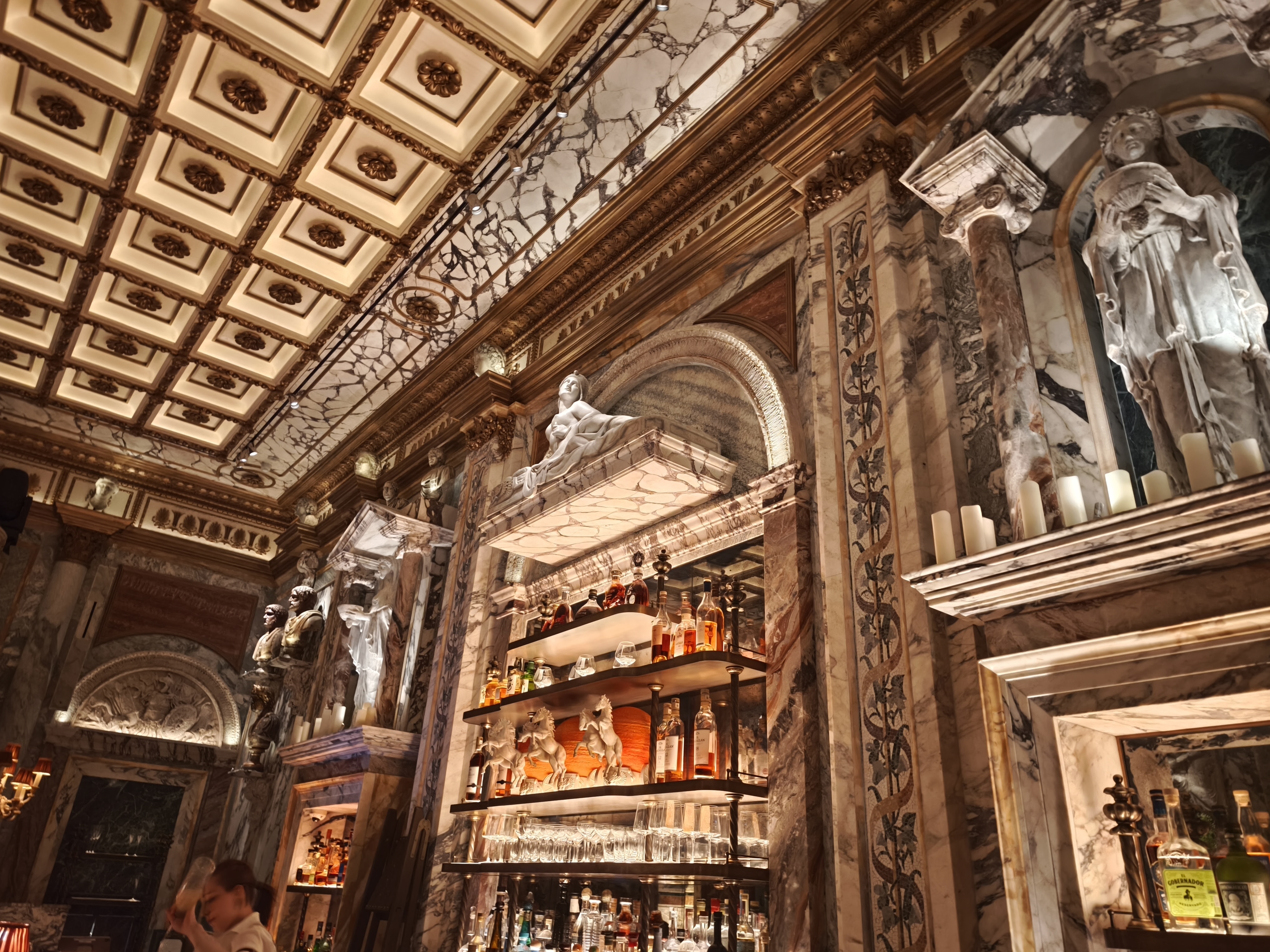 A bar with a lit liquor shelf against a background of marbled walls and a detailed, ornate ceiling with gold trim.