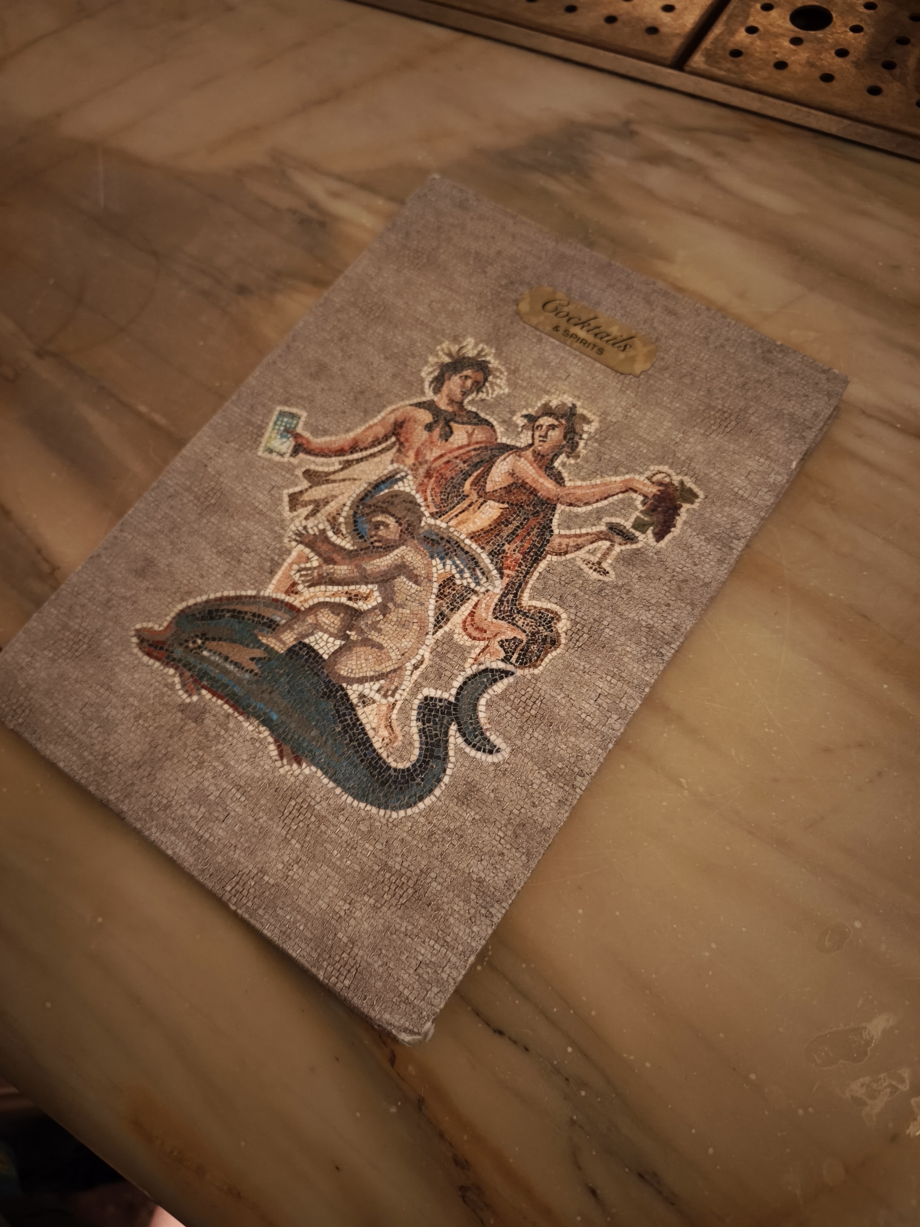 A fabric-covered drinks menu with a colorful mosaic-style illustration of mythological figures on a marble countertop.