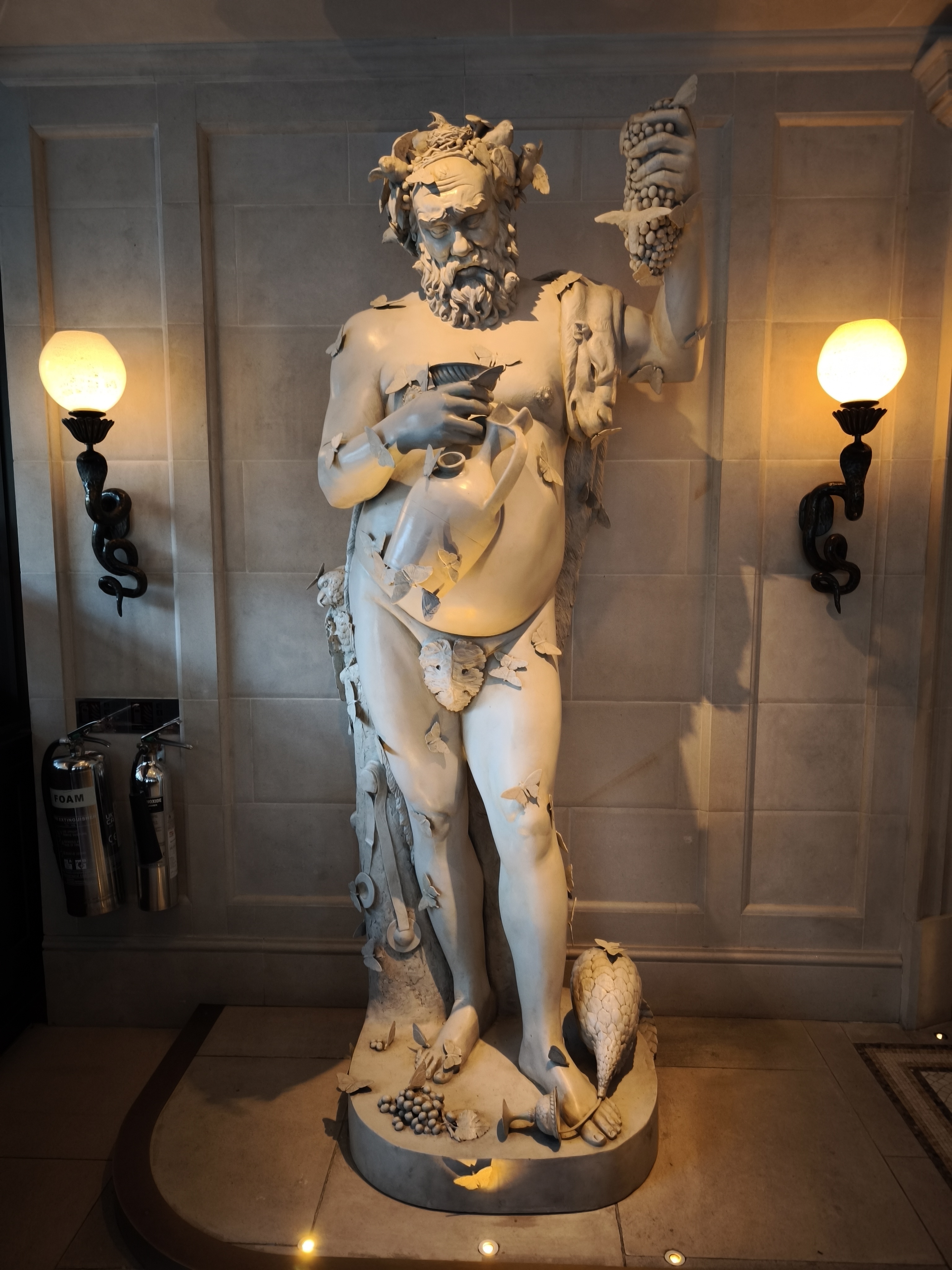 A large, white statue of Bacchus, the Roman god of wine, holding a chalice and grapes, standing in a restaurant entrance with wall sconces on either side.