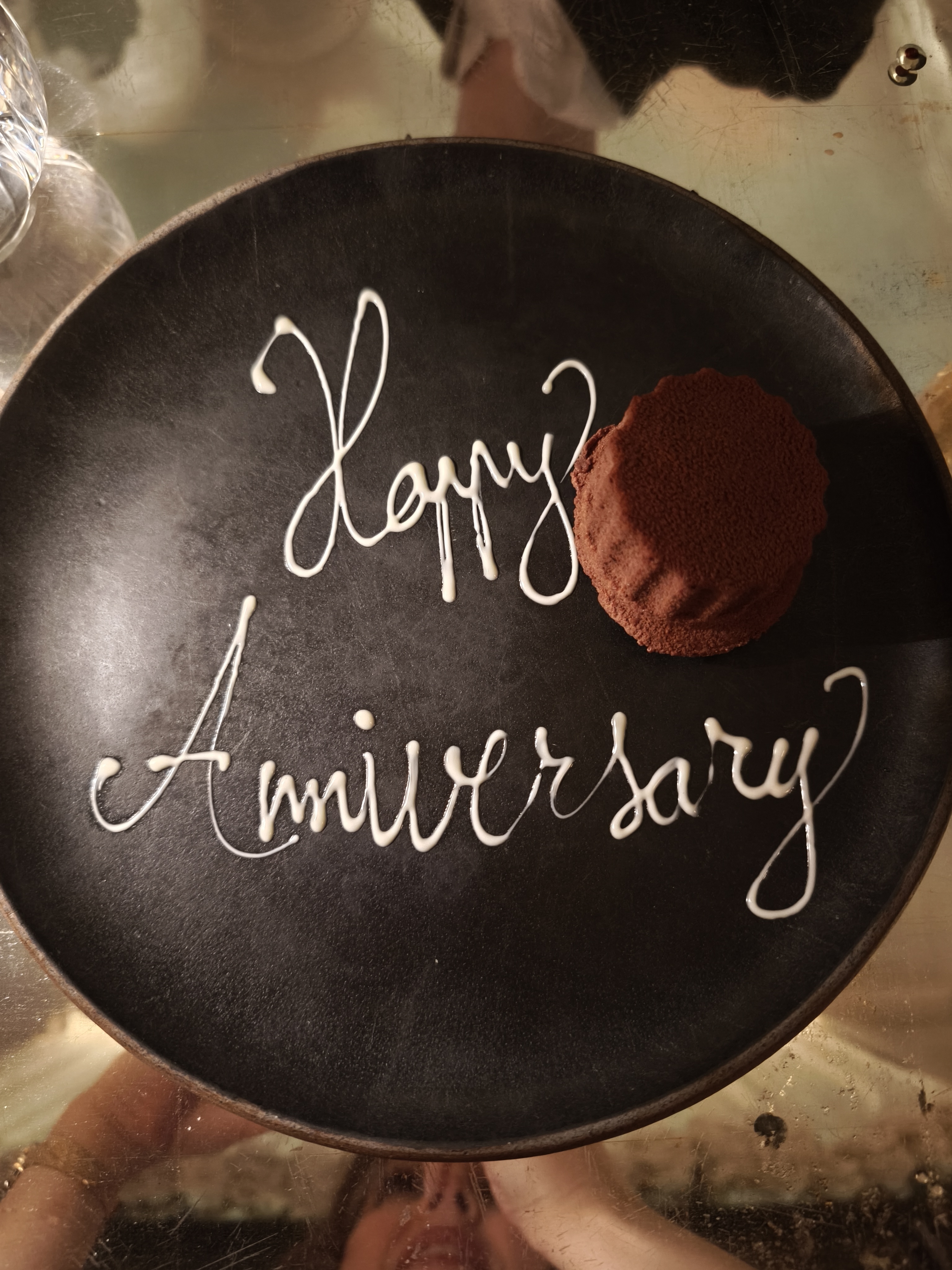 A dark chocolate mousse shaped like a cylinder on a black plate, next to a white chocolate drizzle that spells "Happy Anniversary."