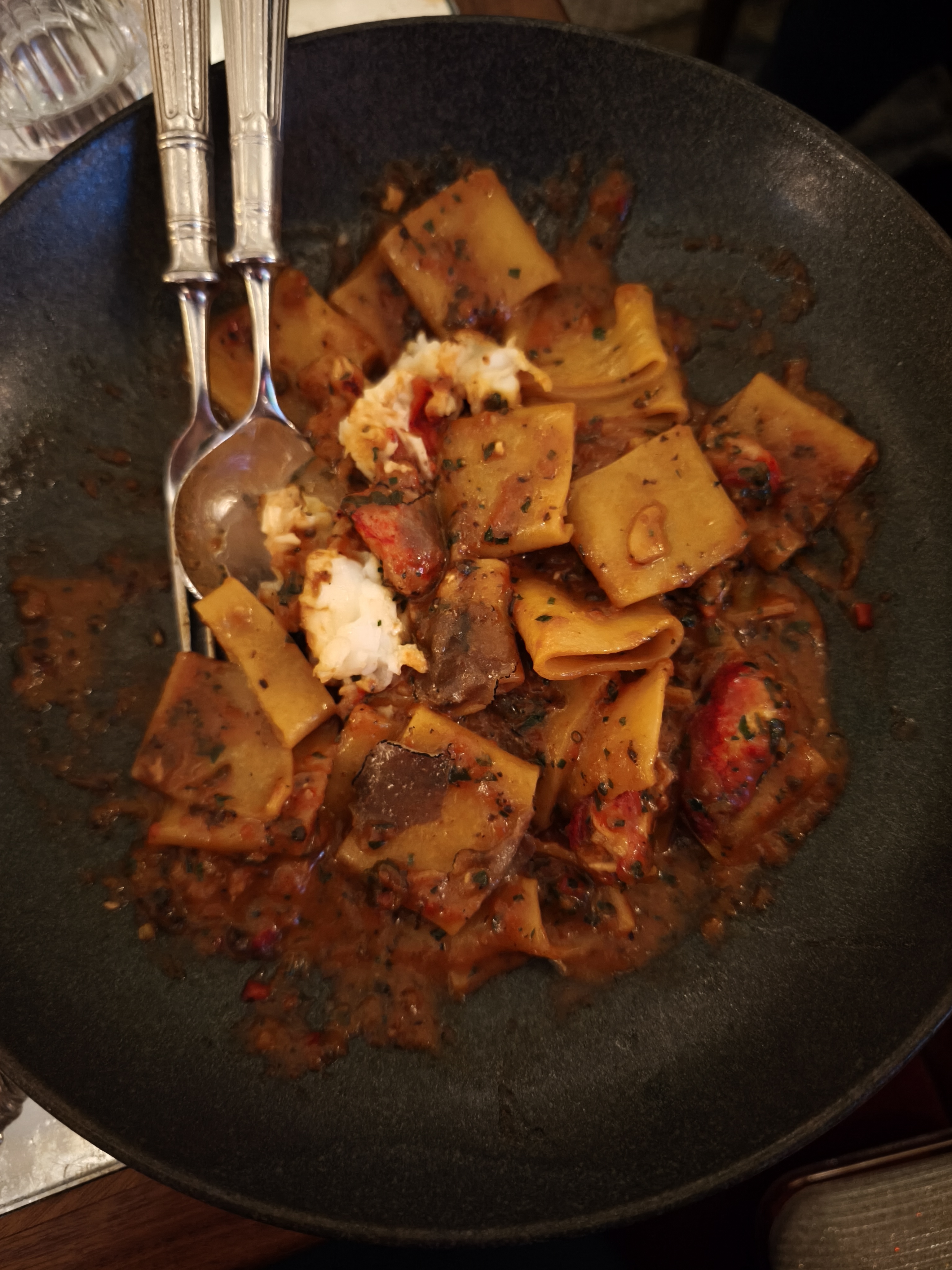 A dark bowl of paccheri pasta with lobster meat and a rich red sauce, next to a fork and spoon.