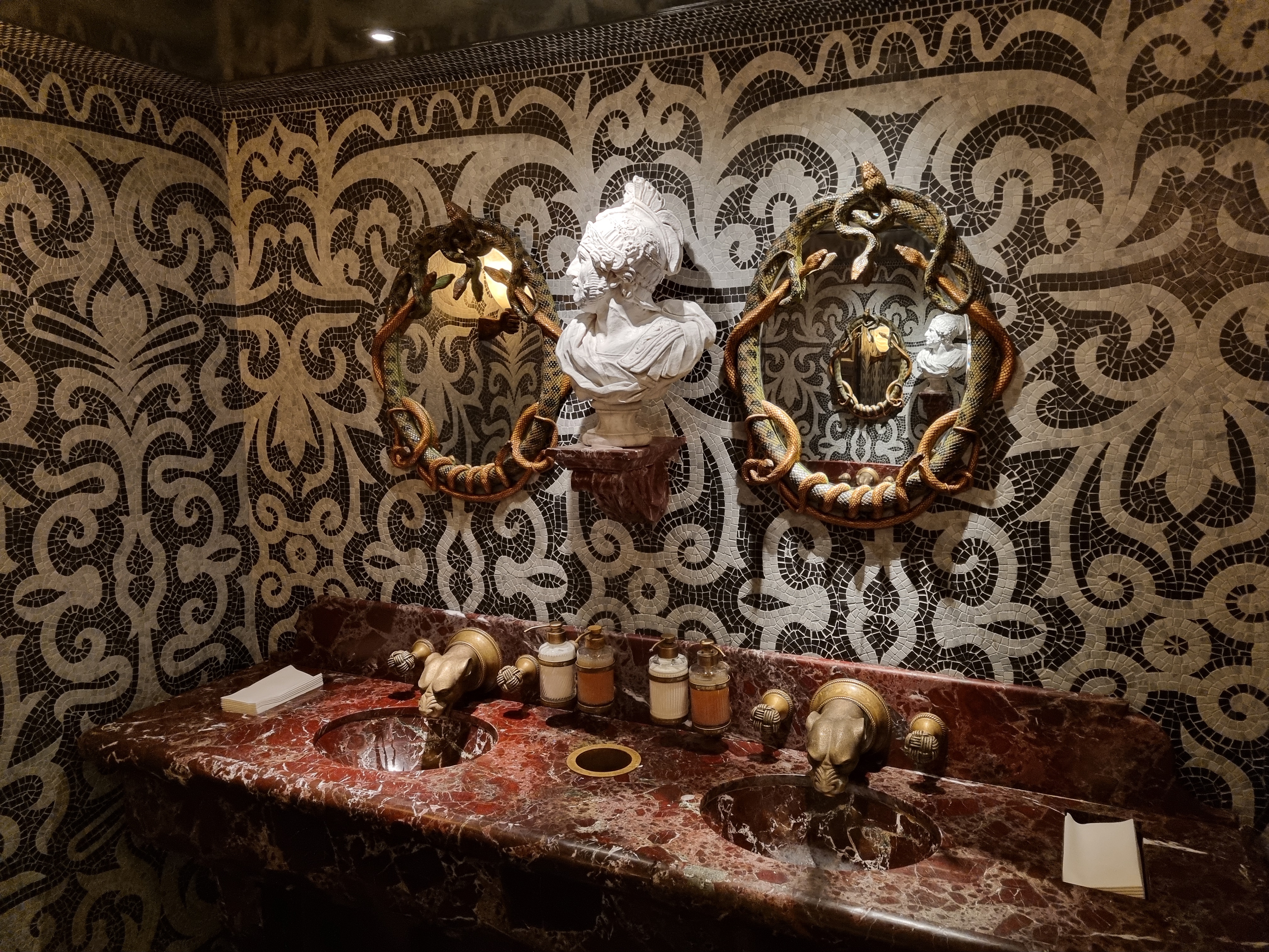A lavishly decorated men's restroom with a red marble double sink, serpent-framed mirrors, and walls covered in dark and light mosaic tiles.