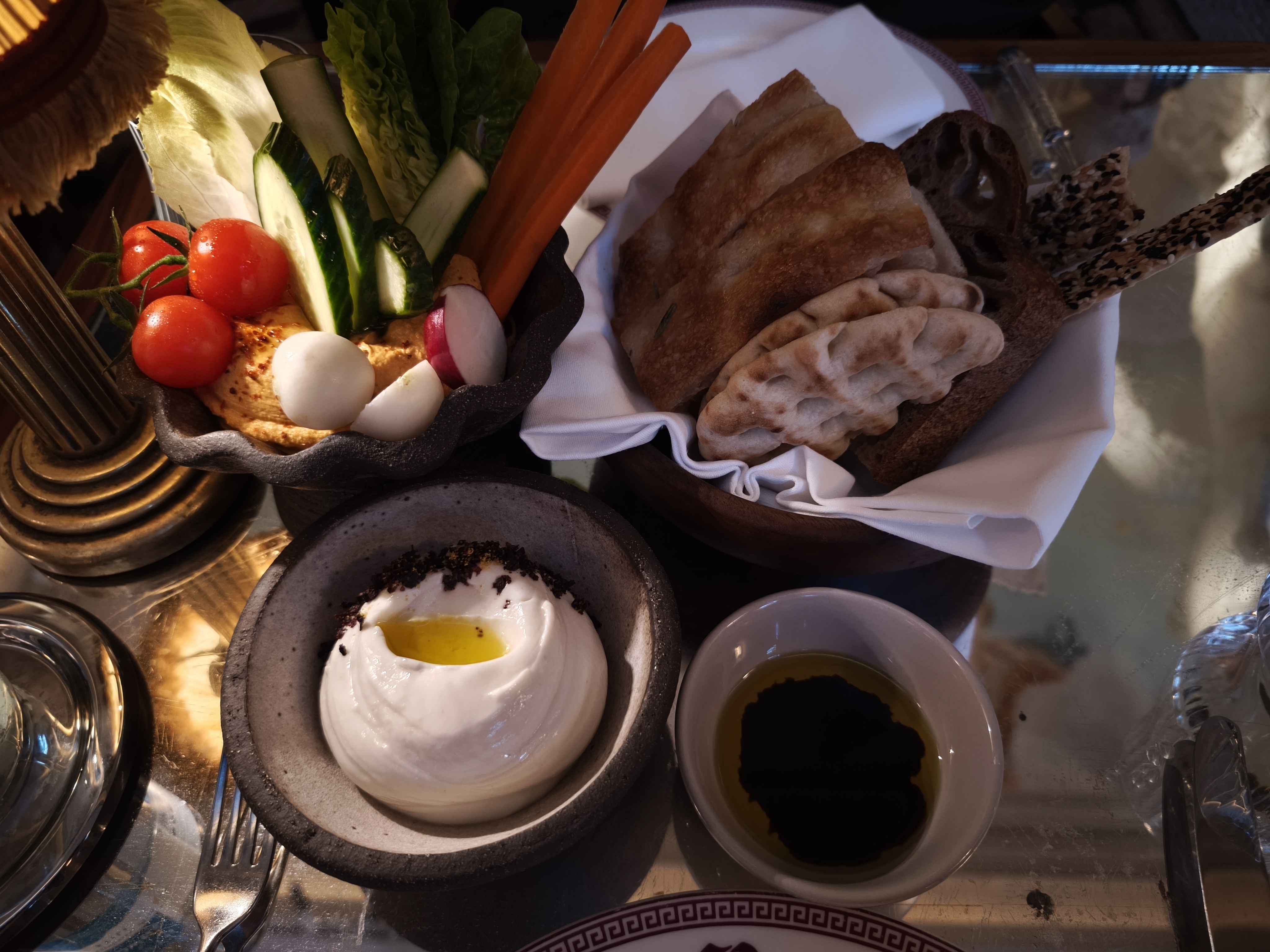A variety of sharing entrees including fresh bread, sliced vegetables, and two bowls of dips. One dip is white with oil and the other is a reddish-orange hummus.