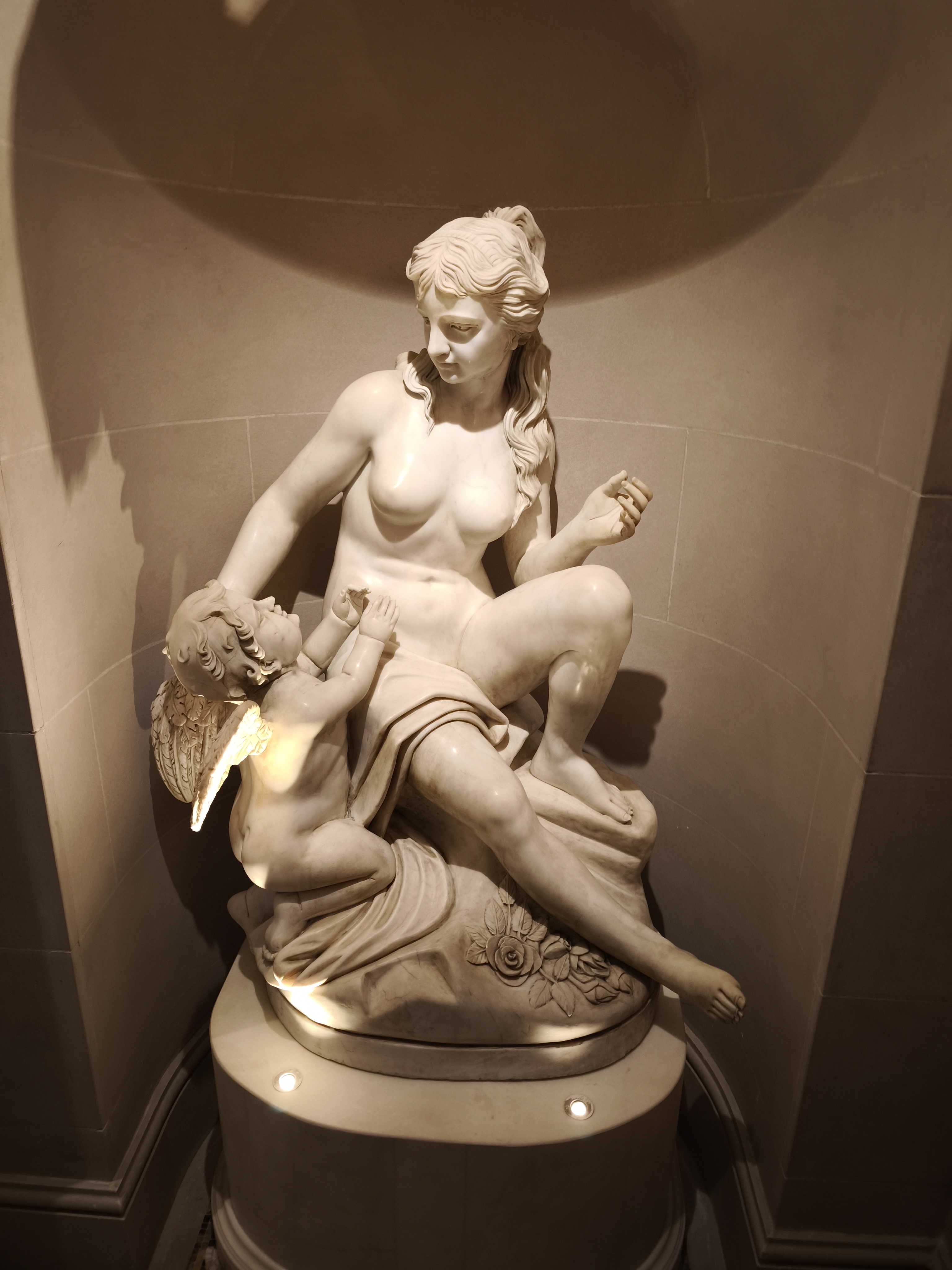 A marble statue of a nude woman seated with a winged baby, illuminated by spotlights in a niche