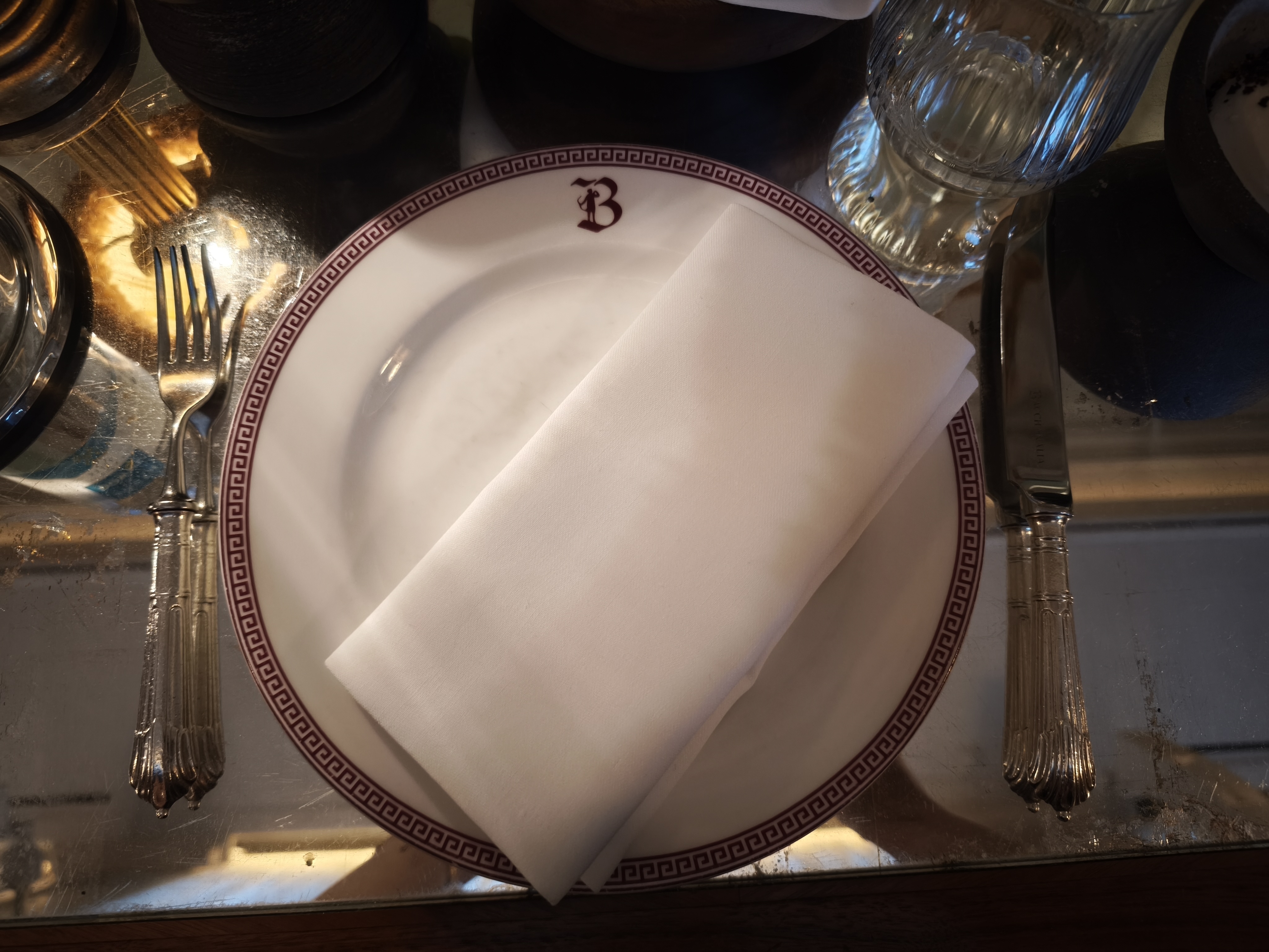 A place setting with a white napkin folded on a white plate with a red Greek key border and a "B" monogram.
