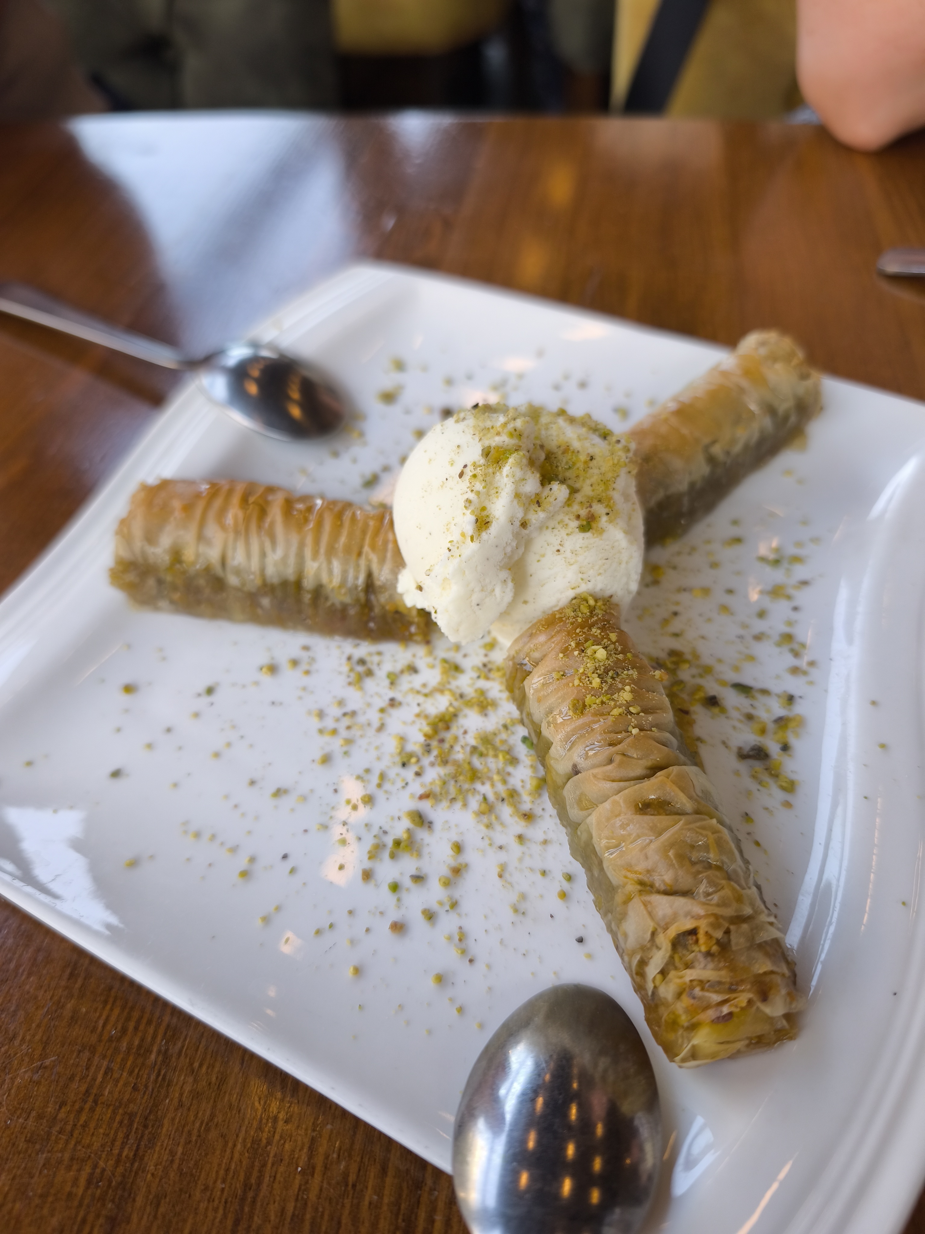 Three pieces of flaky, golden-brown baklava arranged around a scoop of vanilla ice cream, sprinkled with crushed pistachios, on a white plate.