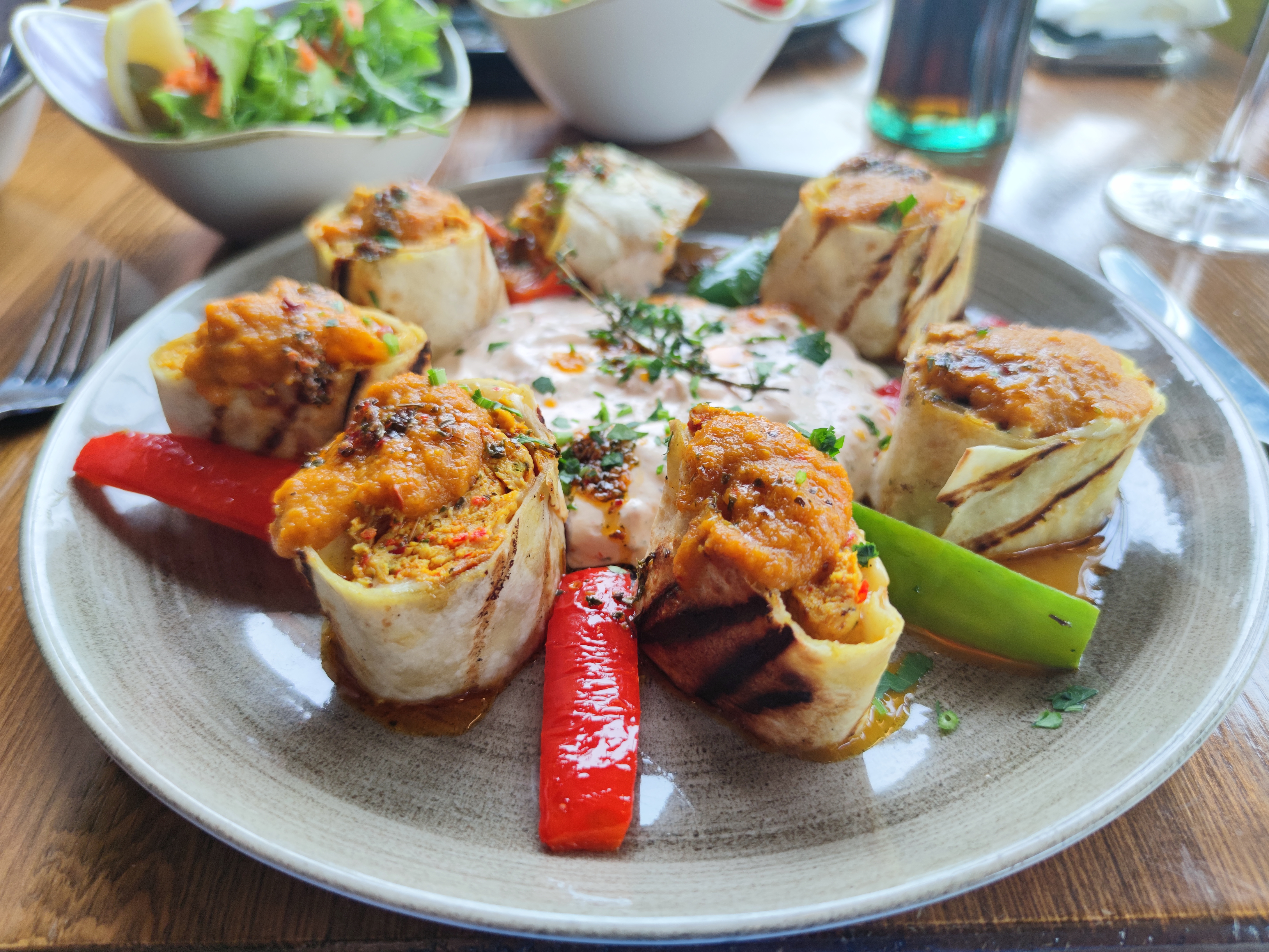 A plate with six rolls of Chicken Beyti arranged around a creamy dip, garnished with grilled red and green peppers and a side salad.