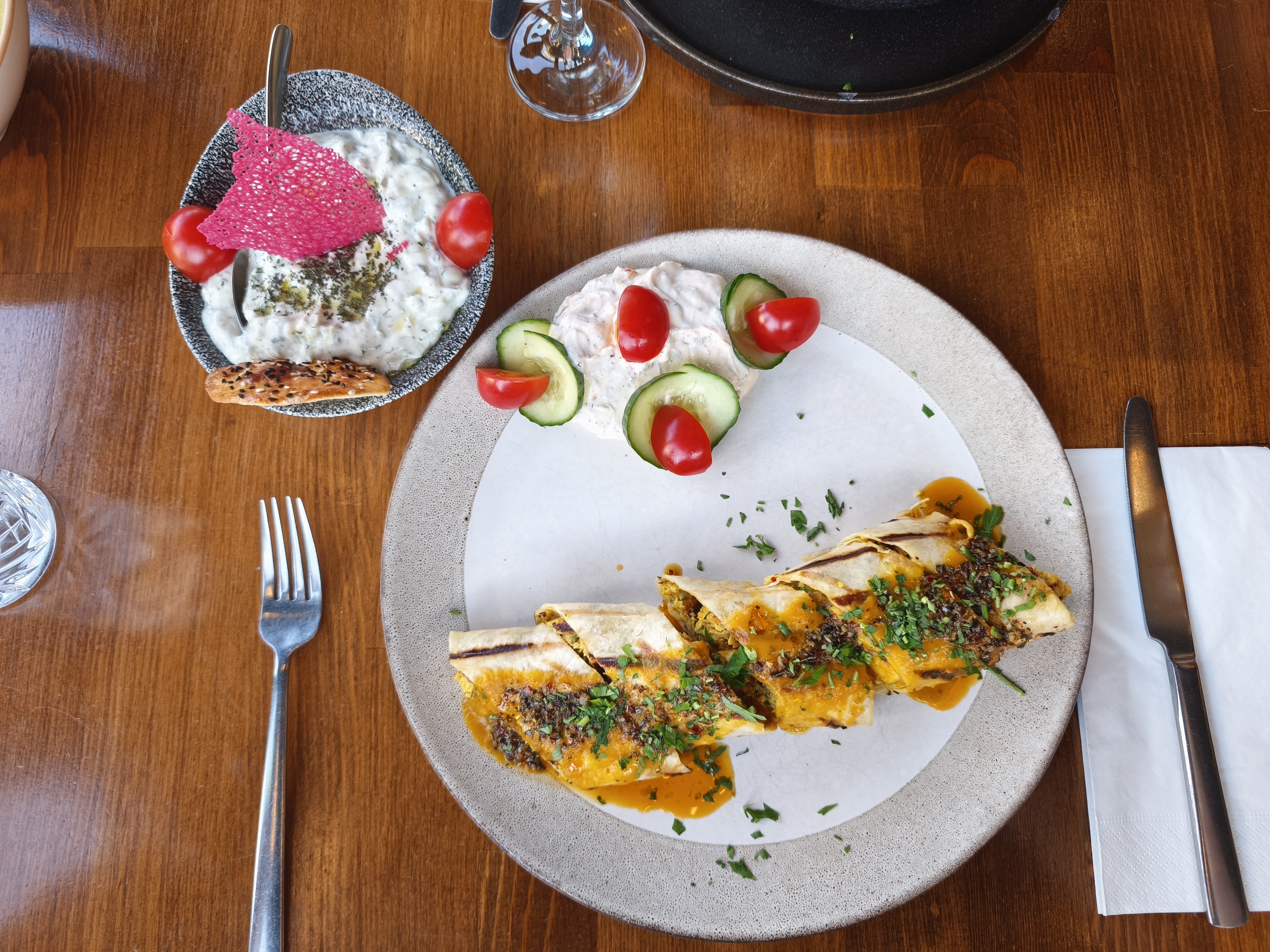 A wide shot of a serving of Chicken Beyti (grilled chicken wrapped in flatbread with sauce) next to a bowl of Tzatziki and a small side of white sauce with cucumbers and tomatoes.