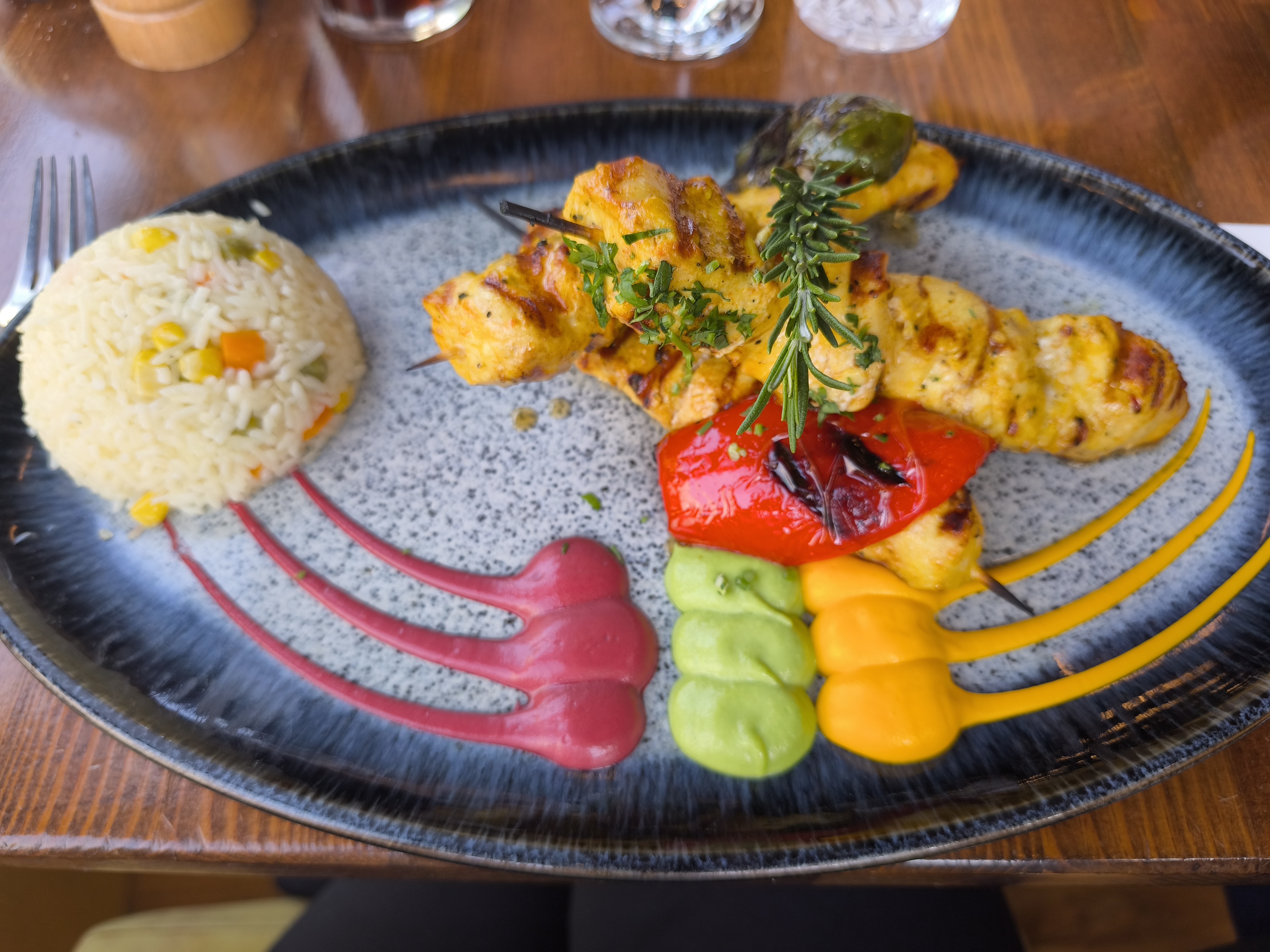 A platter with two chicken skewers and a side of rice, garnished with a sprig of rosemary and colorful sauce swirls.