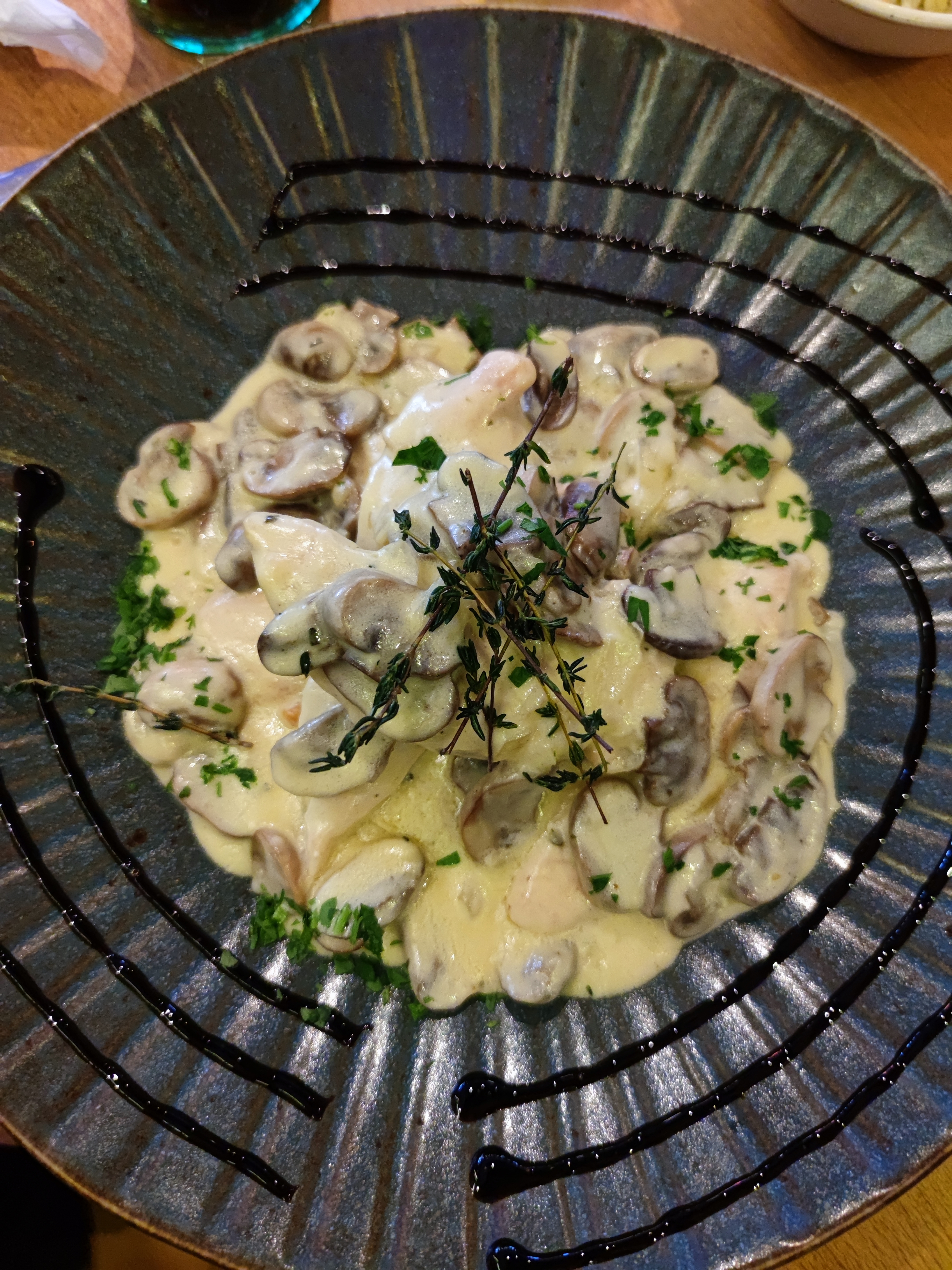 A close-up of a creamy chicken dish topped with mushrooms and fresh herbs, served on a dark blue plate with black sauce drizzles in a circular pattern.