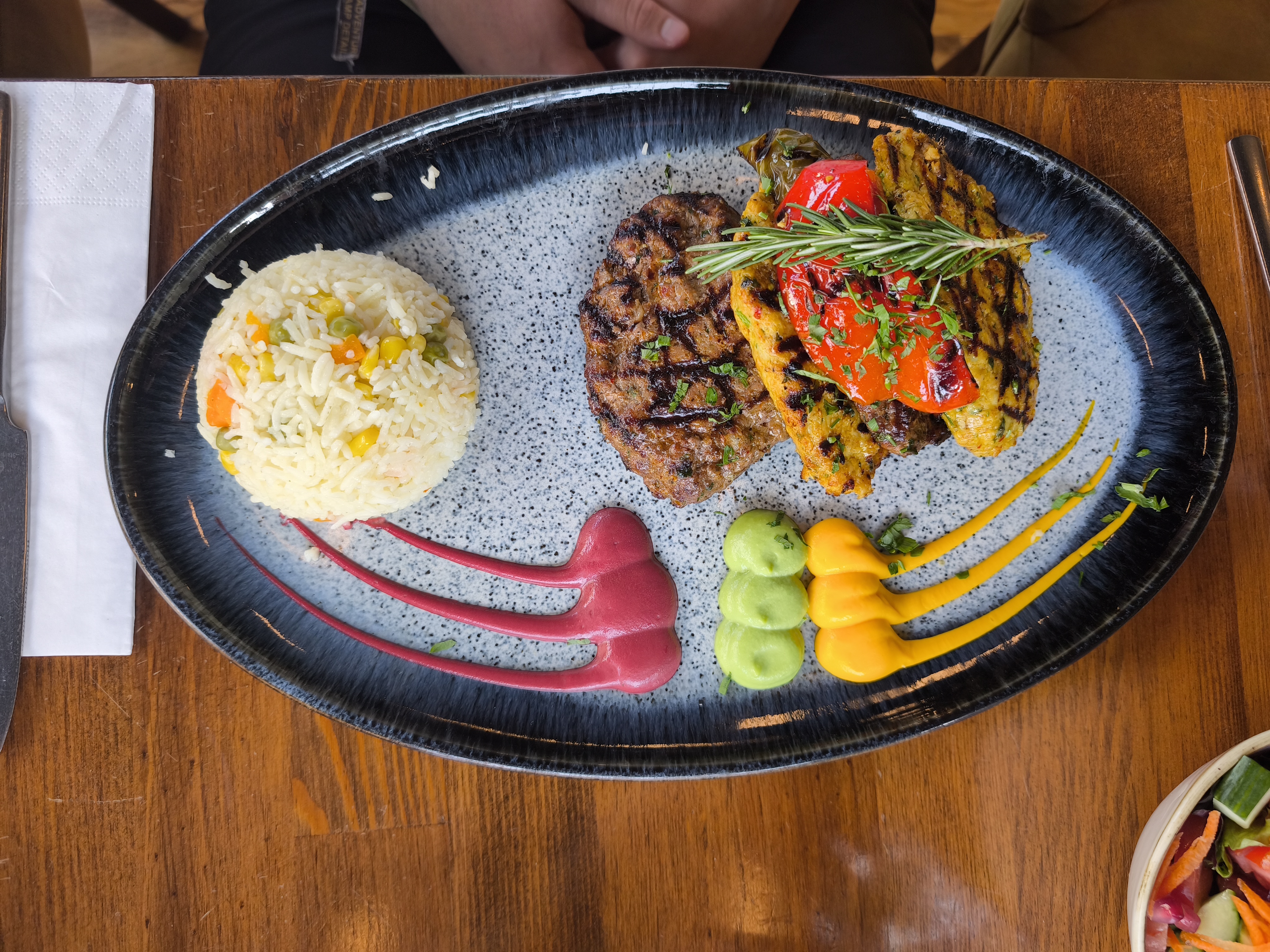 A platter with a grilled meat patty, a skewer of grilled vegetables, a mound of rice with corn and peas, and colorful sauce swirls on the plate.