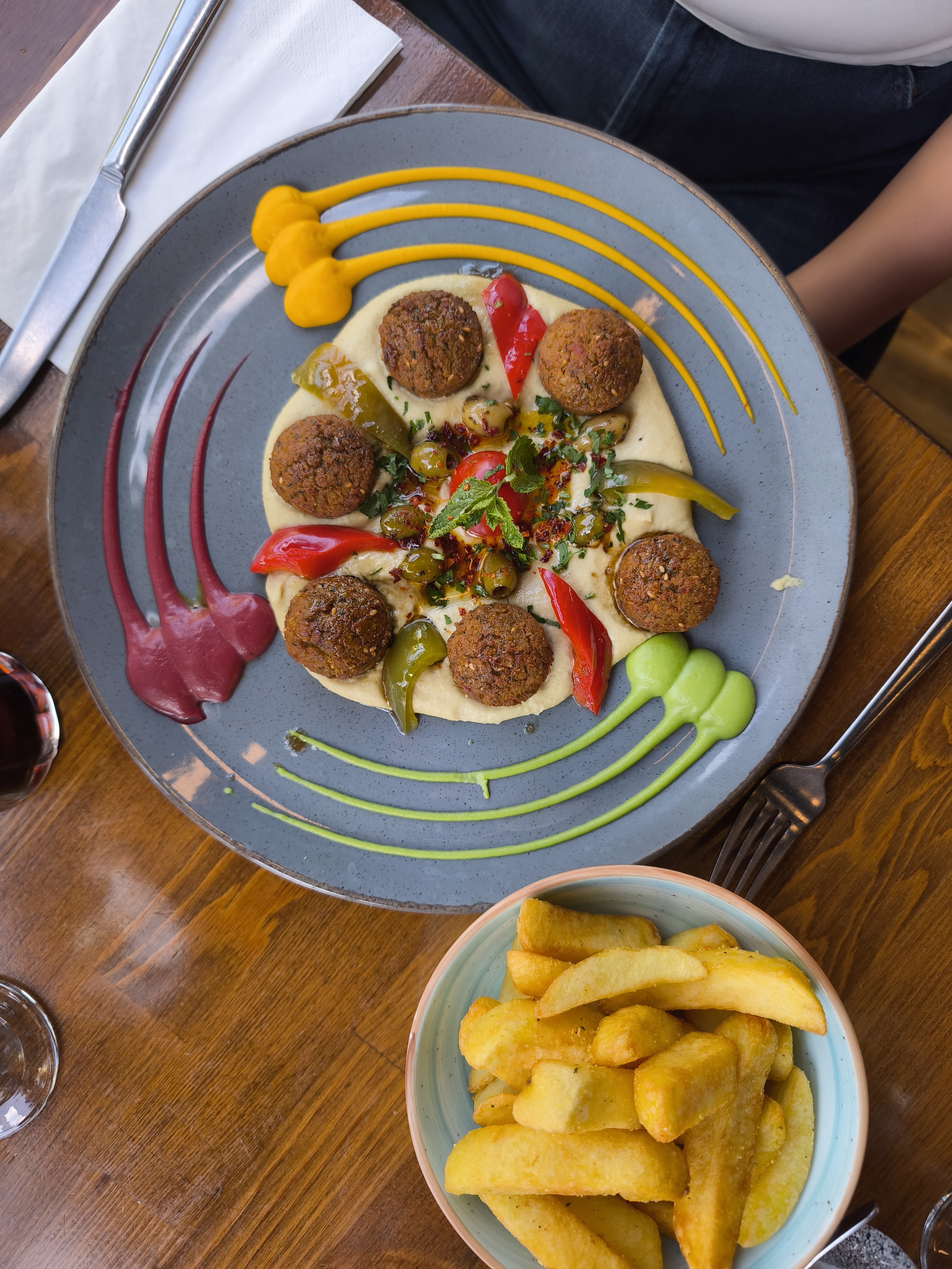 A plate of beautifully arranged falafel balls on a bed of hummus, with colorful sauces and fresh garnishes, accompanied by a bowl of golden chips.