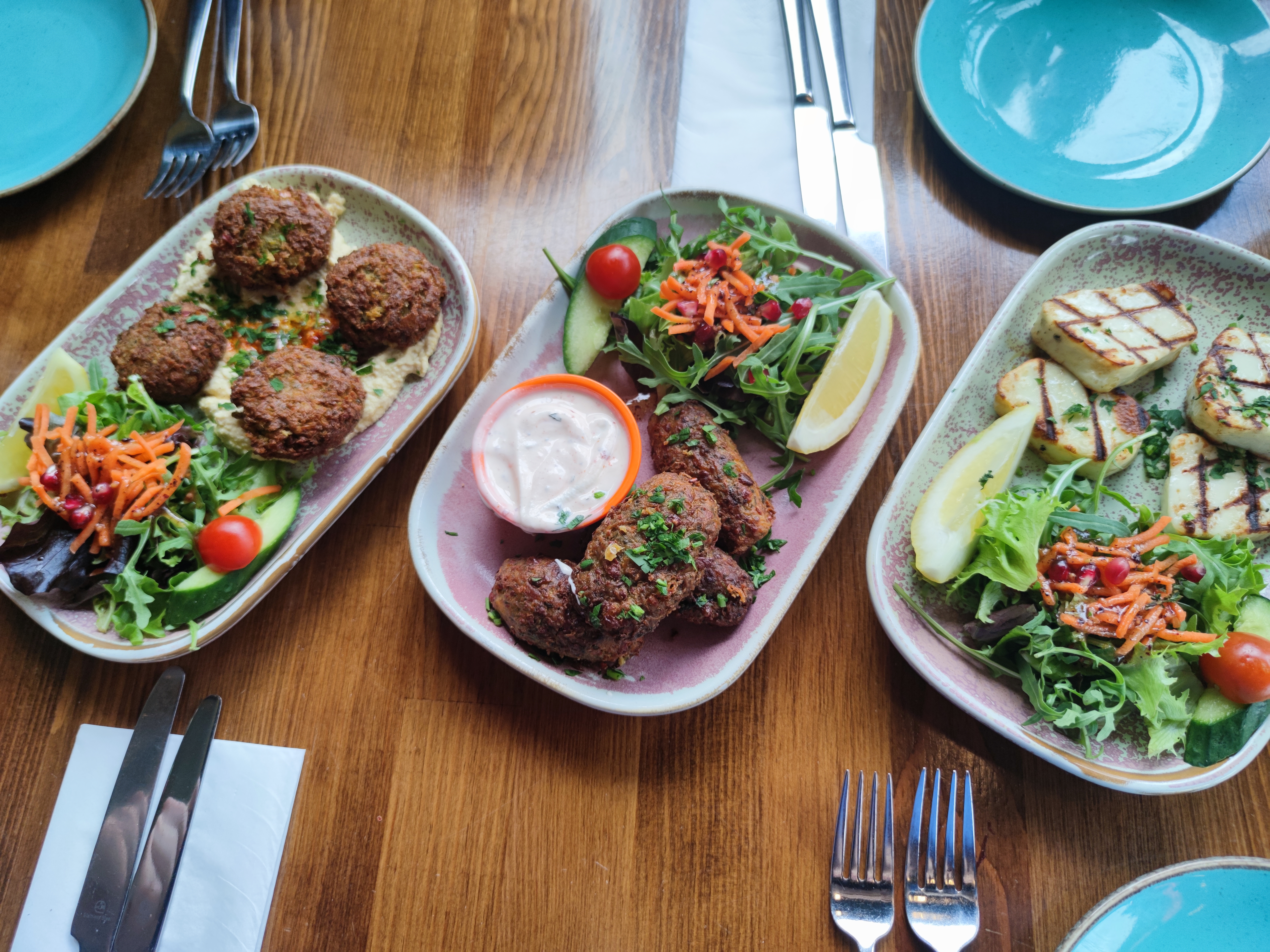 Three appetizer plates on a wooden table: a plate of falafel on hummus, a plate of meat kofte with a yogurt dip, and a plate of grilled halloumi, all with side salads.