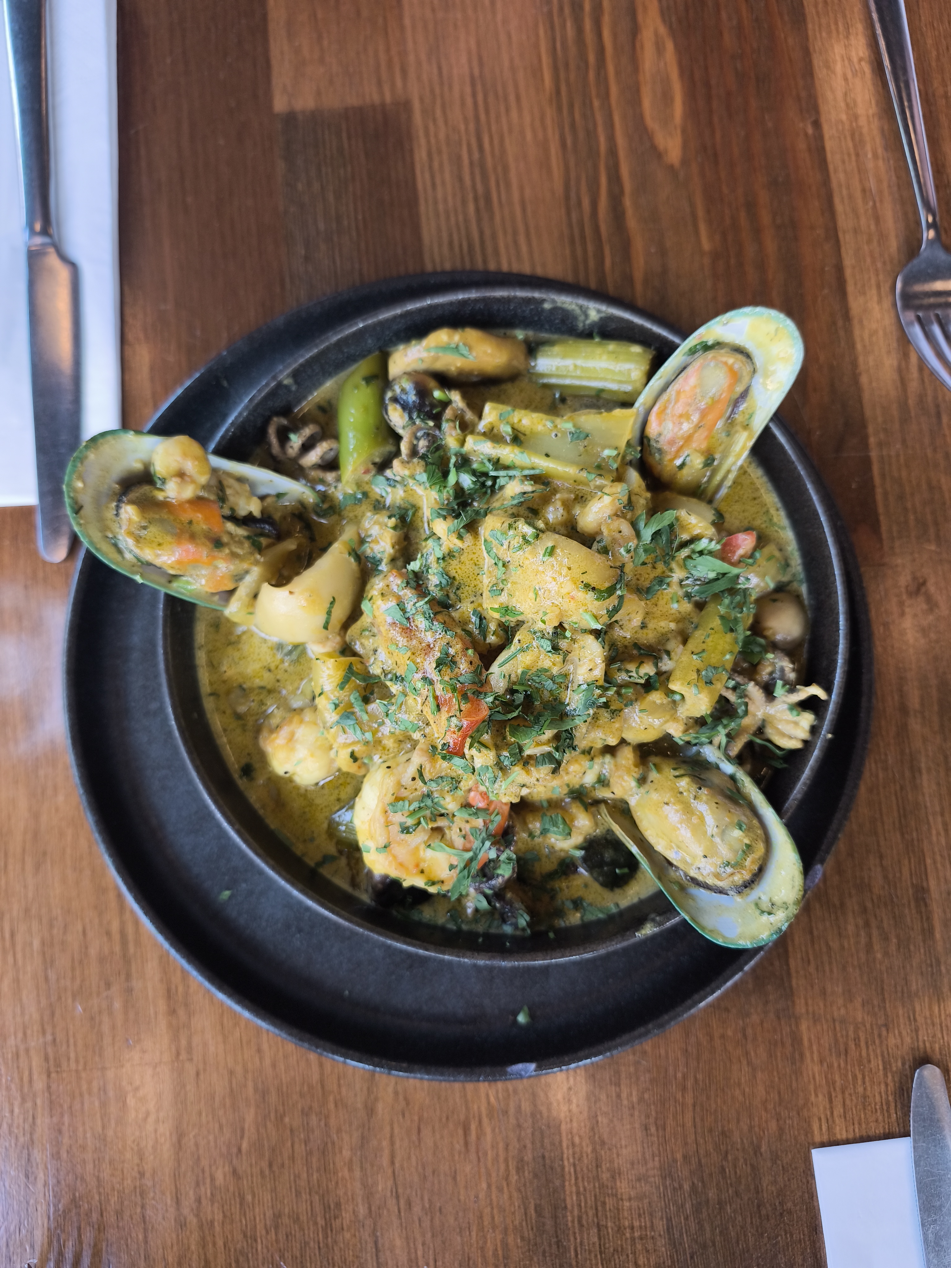 A close-up of a bubbling seafood casserole with large mussels in their shells, calamari, and prawns in a creamy yellow sauce, topped with fresh parsley.