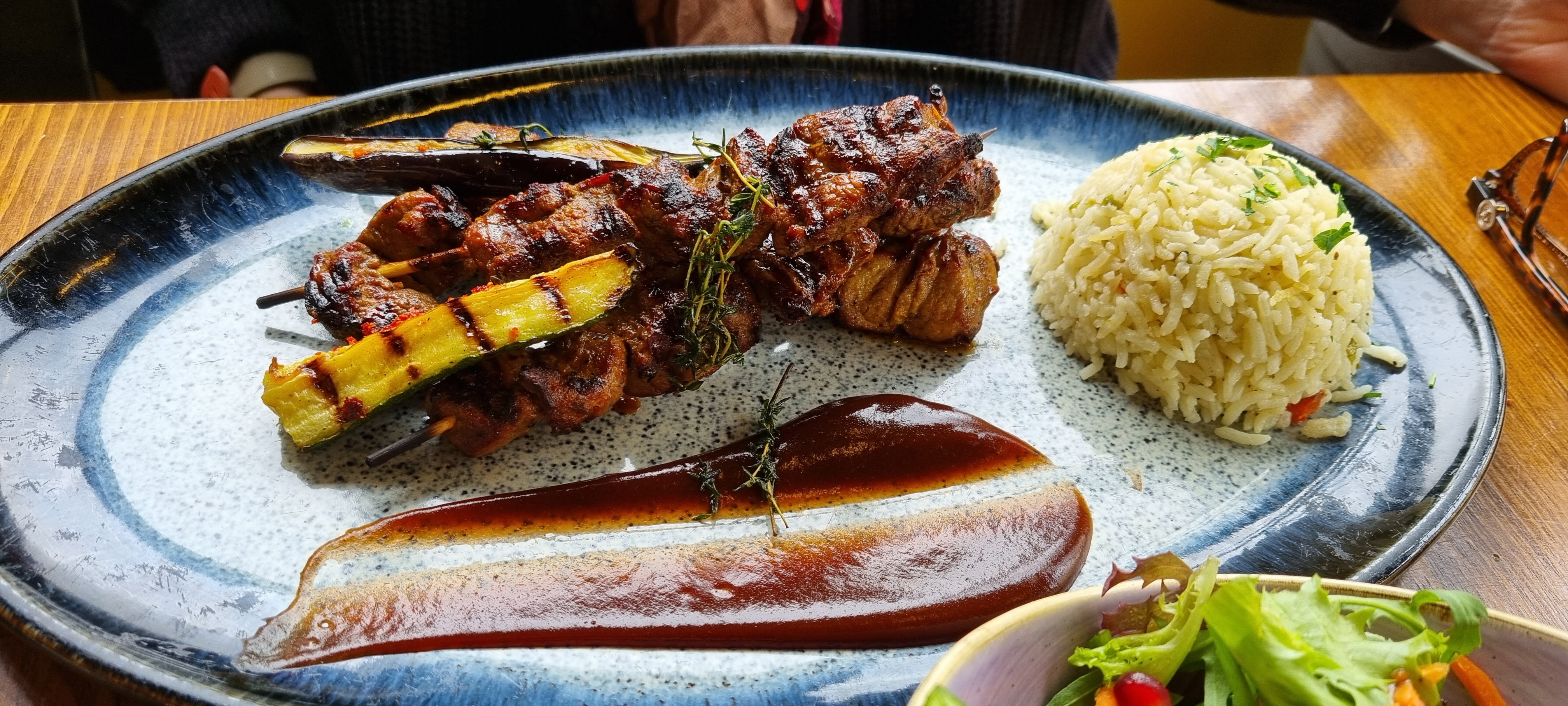 A platter with two skewers of grilled lamb and vegetables, a scoop of rice, and a long piece of dark sauce, presented on a blue and white speckled plate.