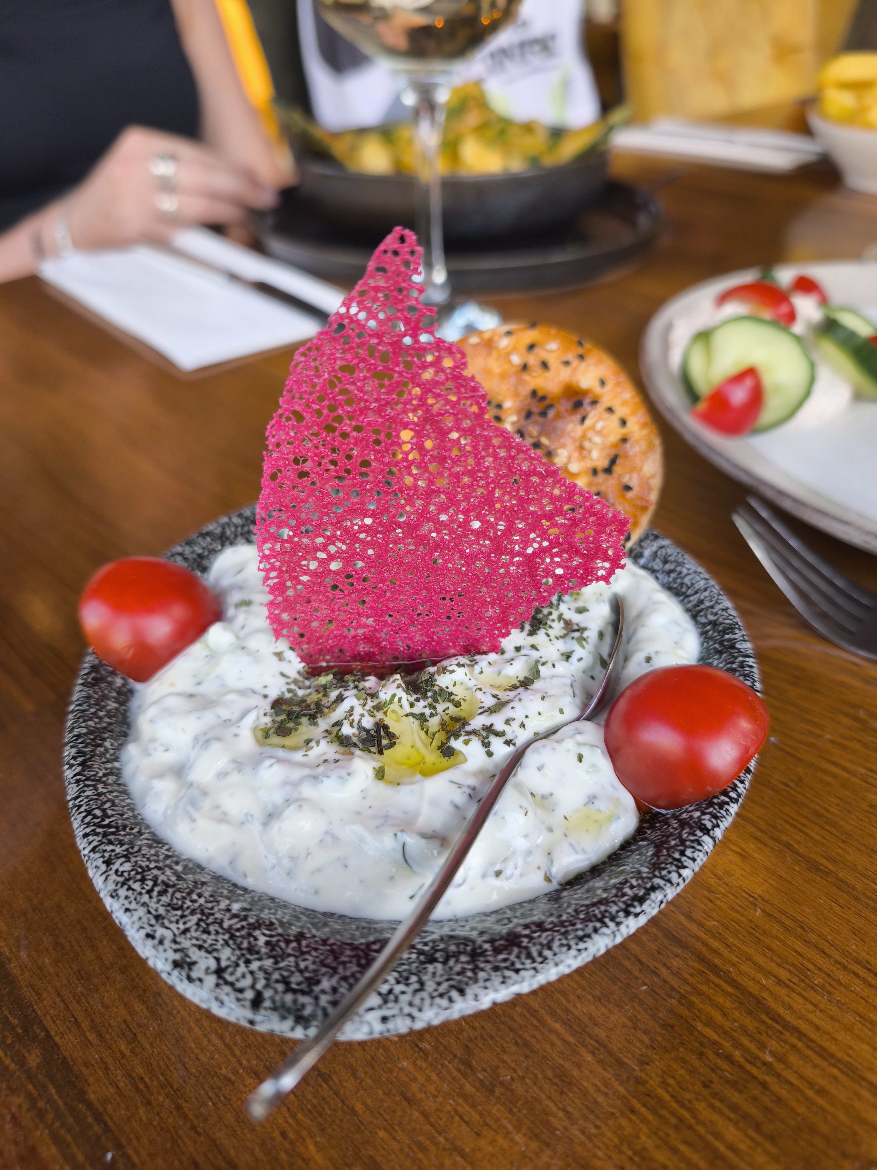 A close-up of a bowl of creamy white Tzatziki with a striking pink crisp, cherry tomatoes, and a drizzle of olive oil.
