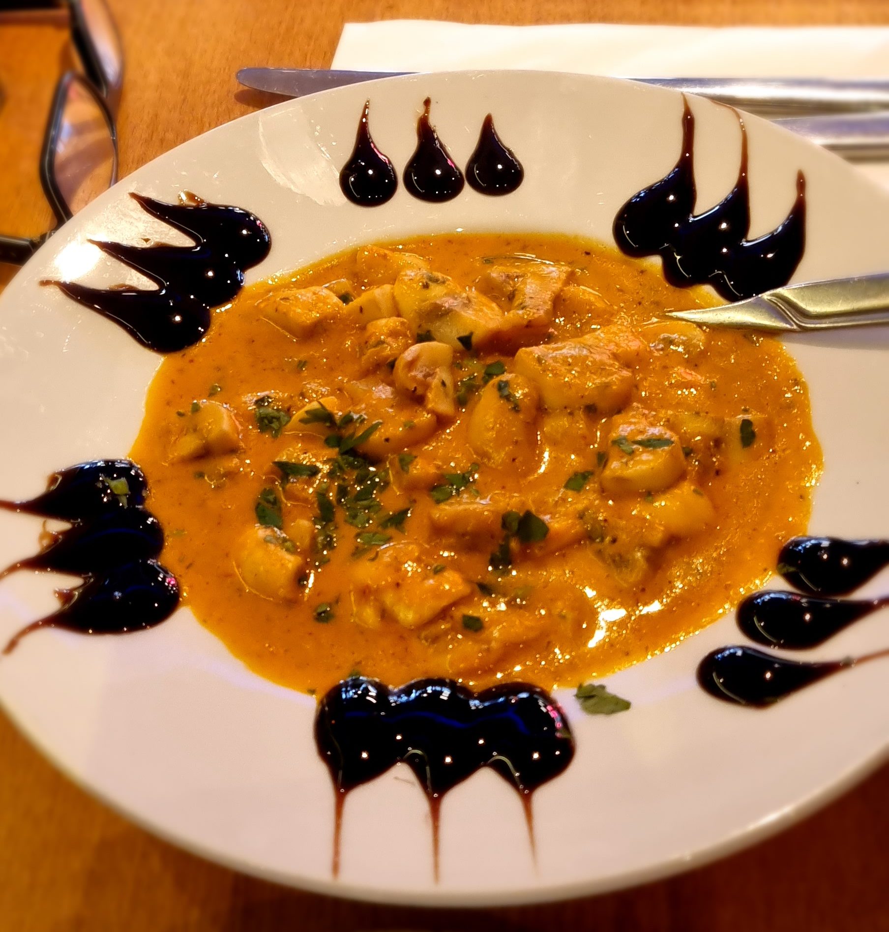 A close-up shot of a warm, orange-colored vegetable casserole with a thick sauce and fresh herbs, served on a white plate with dark drizzles around the rim.
