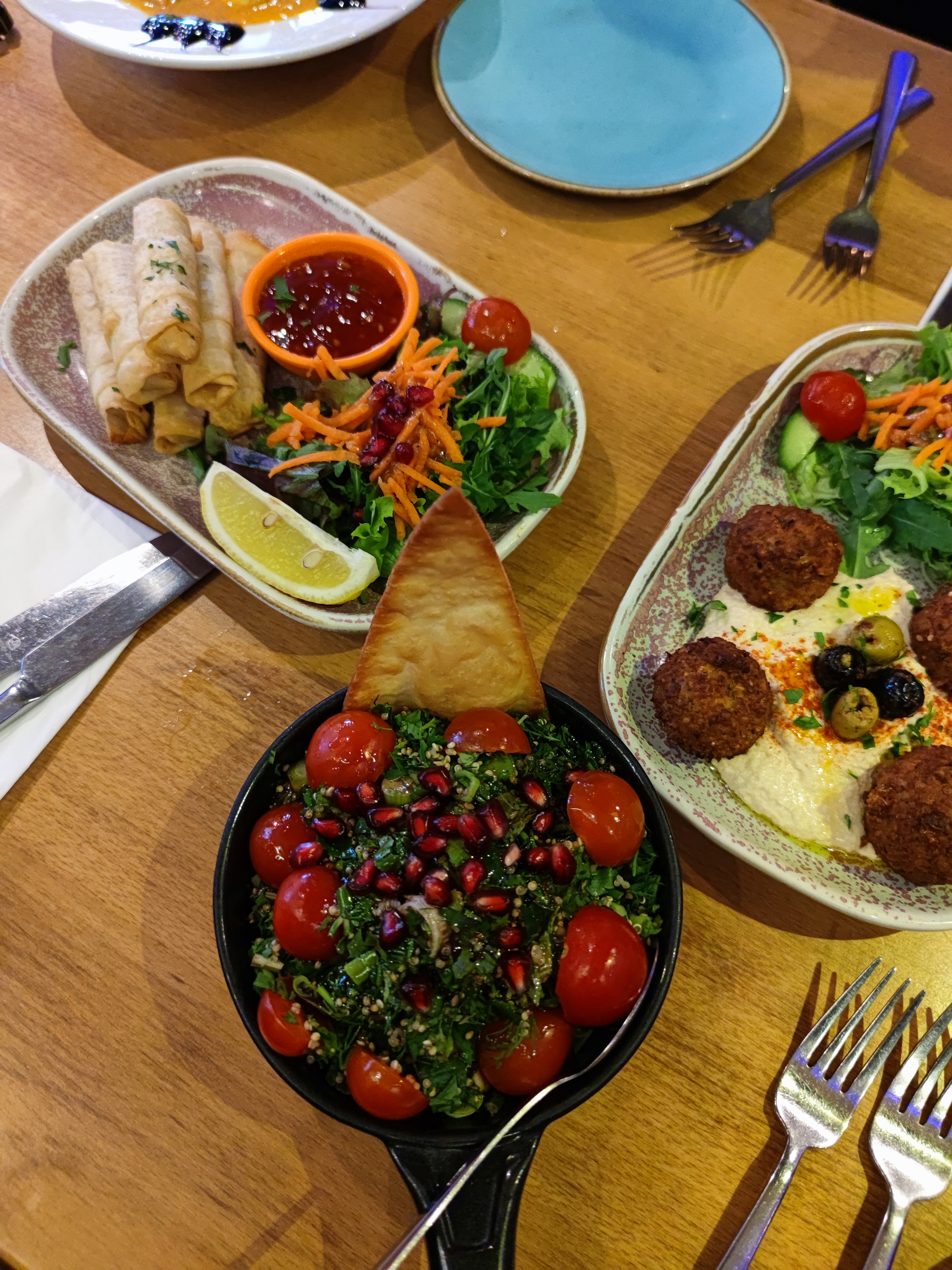 A table with three appetizer plates: a bowl of Tabbouleh salad, a plate of crispy cheese rolls with a dipping sauce, and a plate of falafel on hummus.