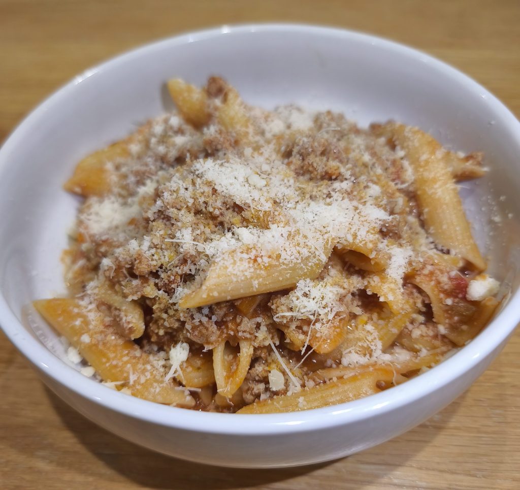 A bowl of penne bolognese topped with a layer of grated cheese.