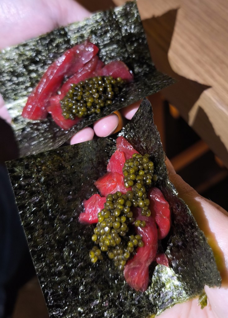 Two hands holding up small square sheets of dark green nori seaweed, each topped with bright red, scraped tuna meat (Nakaochi) and a mound of green-gold N25 Caviar.
