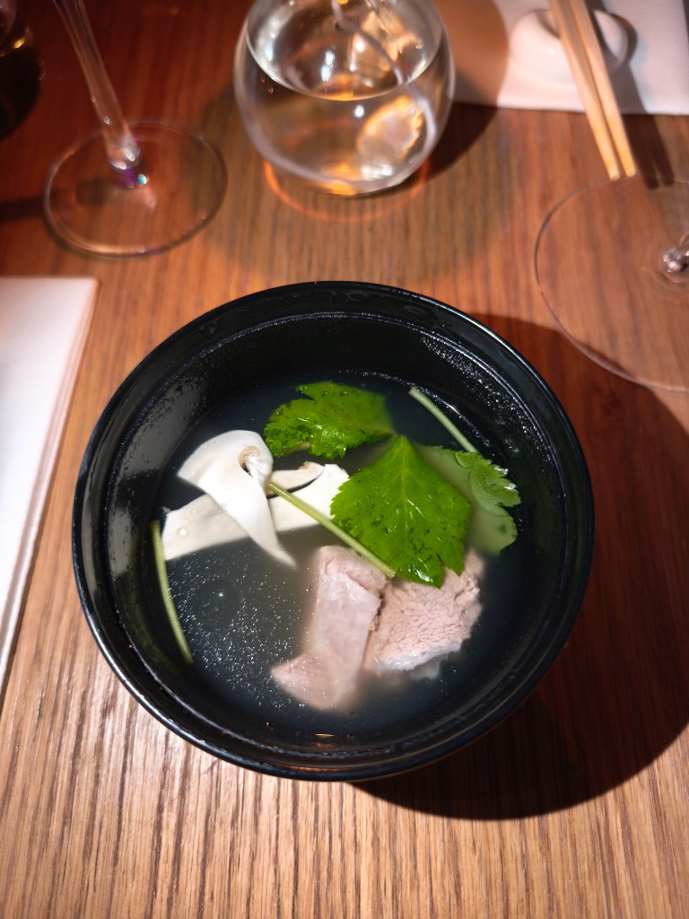 A black bowl containing clear broth with slices of white mushroom, a piece of light pink meat, and a large green leaf garnish.