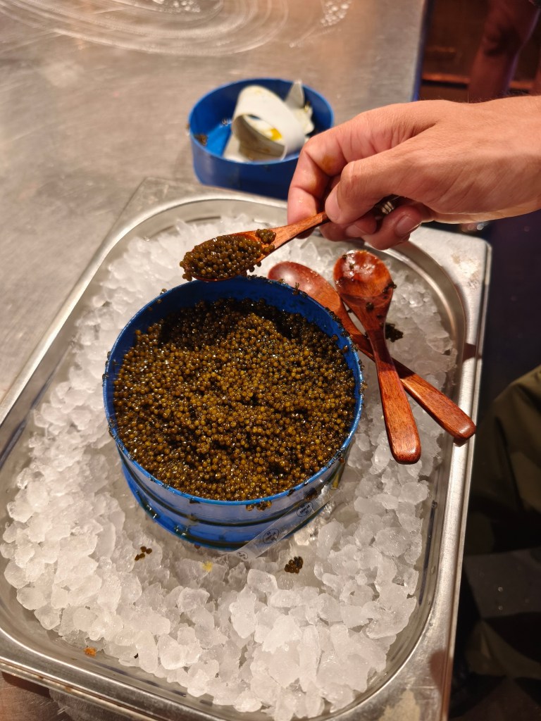 A blue metal tin of deep bronze-coloured N25 caviar sitting on a bed of crushed ice, with a small wooden spoon hovering over it.