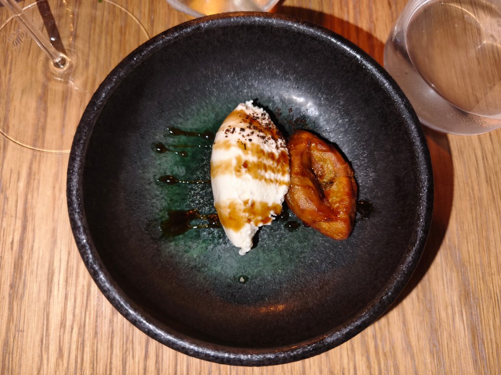 A dark ceramic bowl holding a quenelle of white ice cream drizzled with dark syrup and a caramelized piece of sweet potato.