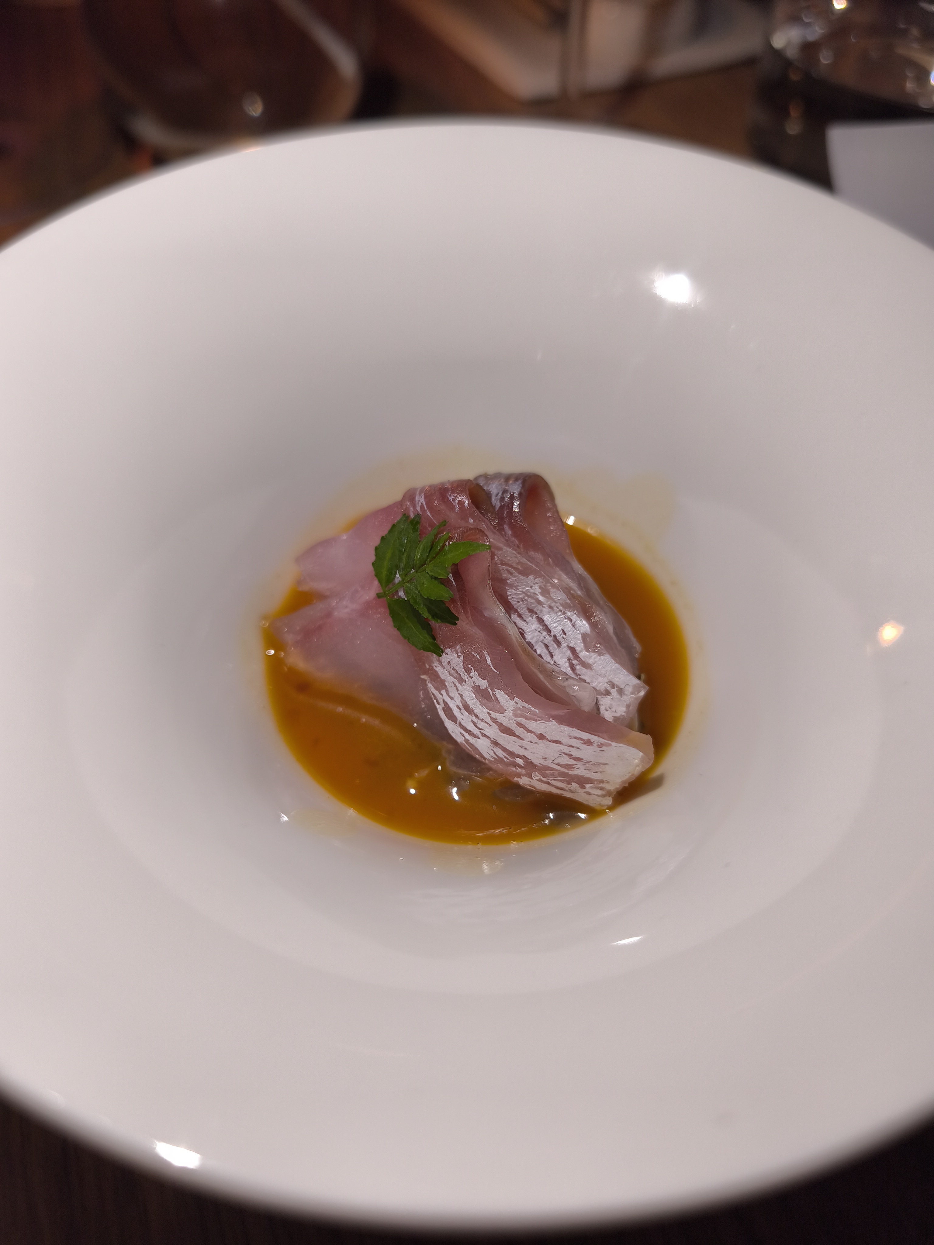 A close-up of a small serving of raw, thinly sliced sea bream with white skin, presented in a deep white bowl with a bright orange-yellow sauce at the bottom.