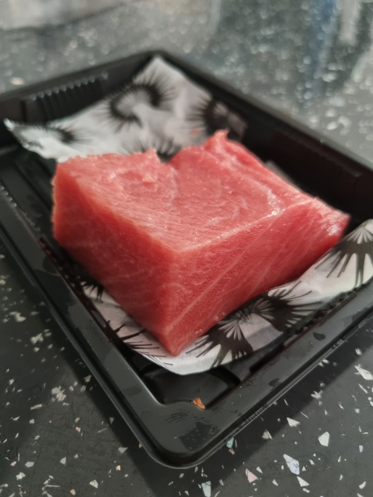 A thick, square cut of raw, bright red Bluefin tuna steak sitting in a black plastic container lined with patterned paper.